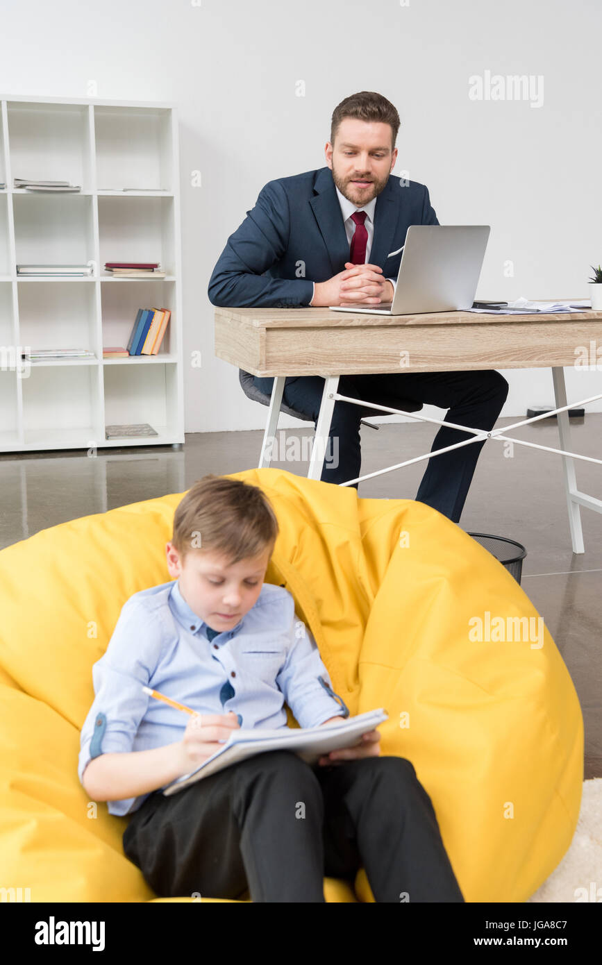 Ragazzo seduto sul cuscino giallo e disegno mentre suo padre imprenditore lavora in ufficio Foto Stock