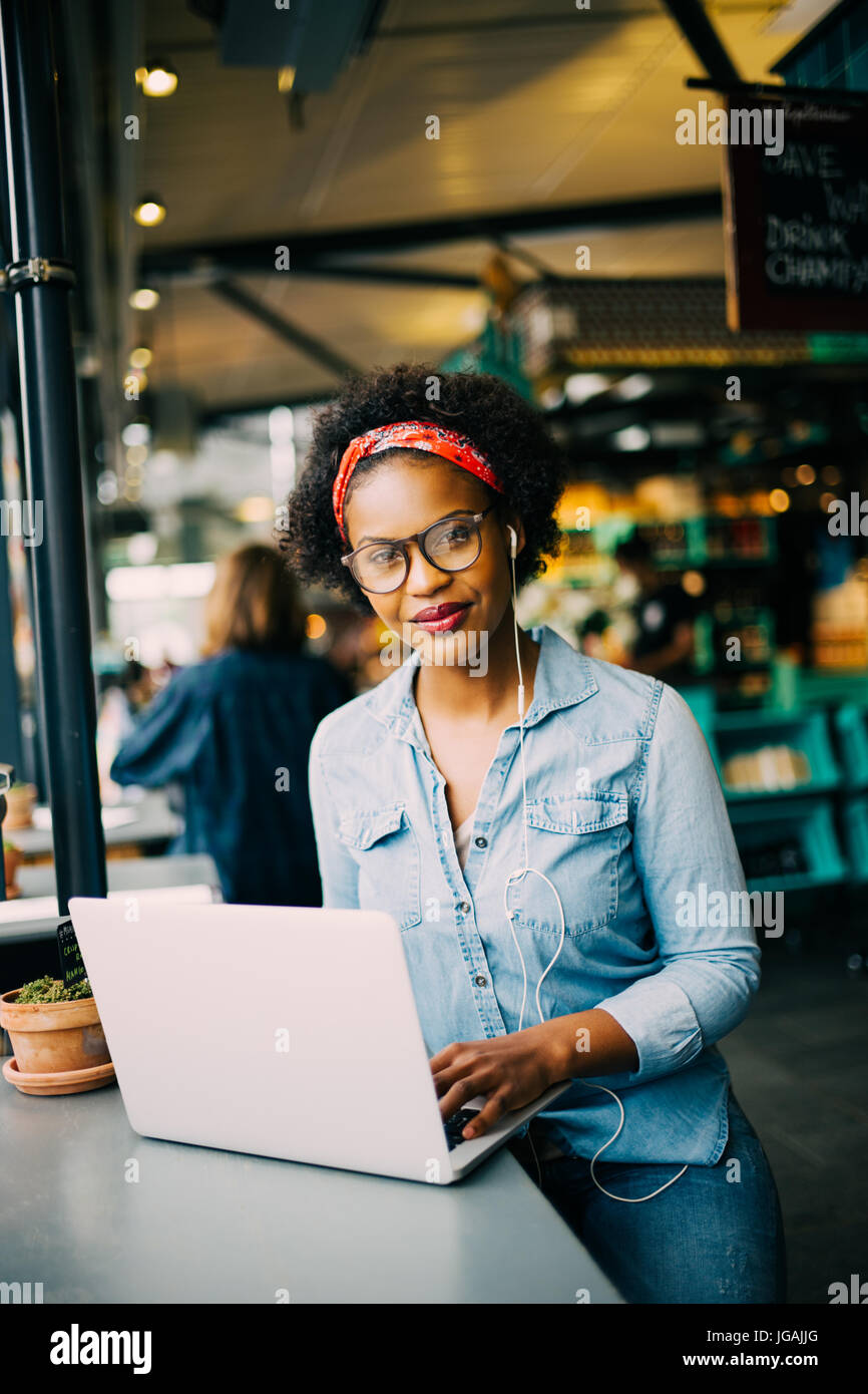 Sorridente giovane donna africana seduto da solo ad un contatore in un bistro lavorando su un computer portatile e di ascoltare la musica in cuffia Foto Stock