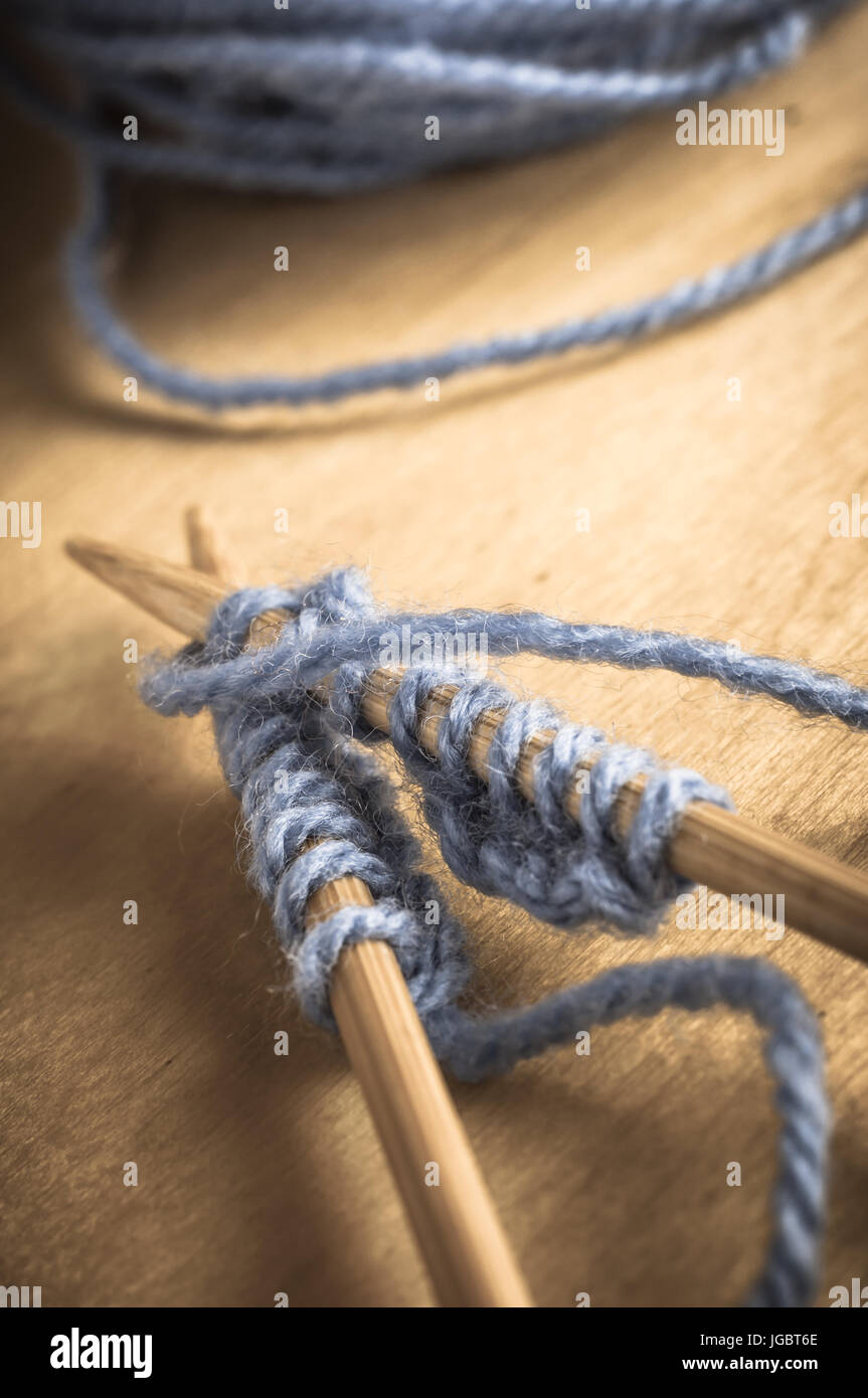 Una coppia incrociata di aghi da maglia sul tavolo di legno contenente punti di cucitura in corso con blu pallido e filati di lana palla in background. Leggermente sbiadite Foto Stock