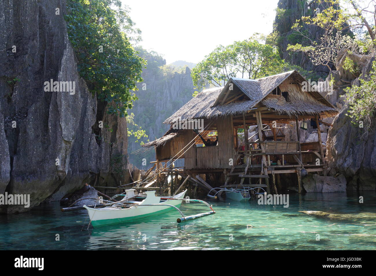 Piccola cabina in legno / capanna su palafitte e rocce, nella splendida laguna blu con ripide scogliere. Uomo in amaca. Le piccole imbarcazioni da pesca. Coron Filippine. Foto Stock
