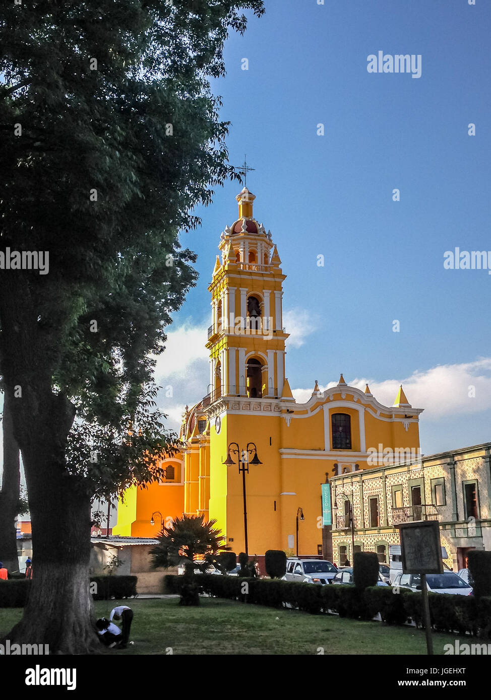 Di colore giallo brillante facciata di San Apostolo chiesa nel Zocalo in Cholula, Messico visto attraverso prati e arbusti contro un cielo blu Foto Stock