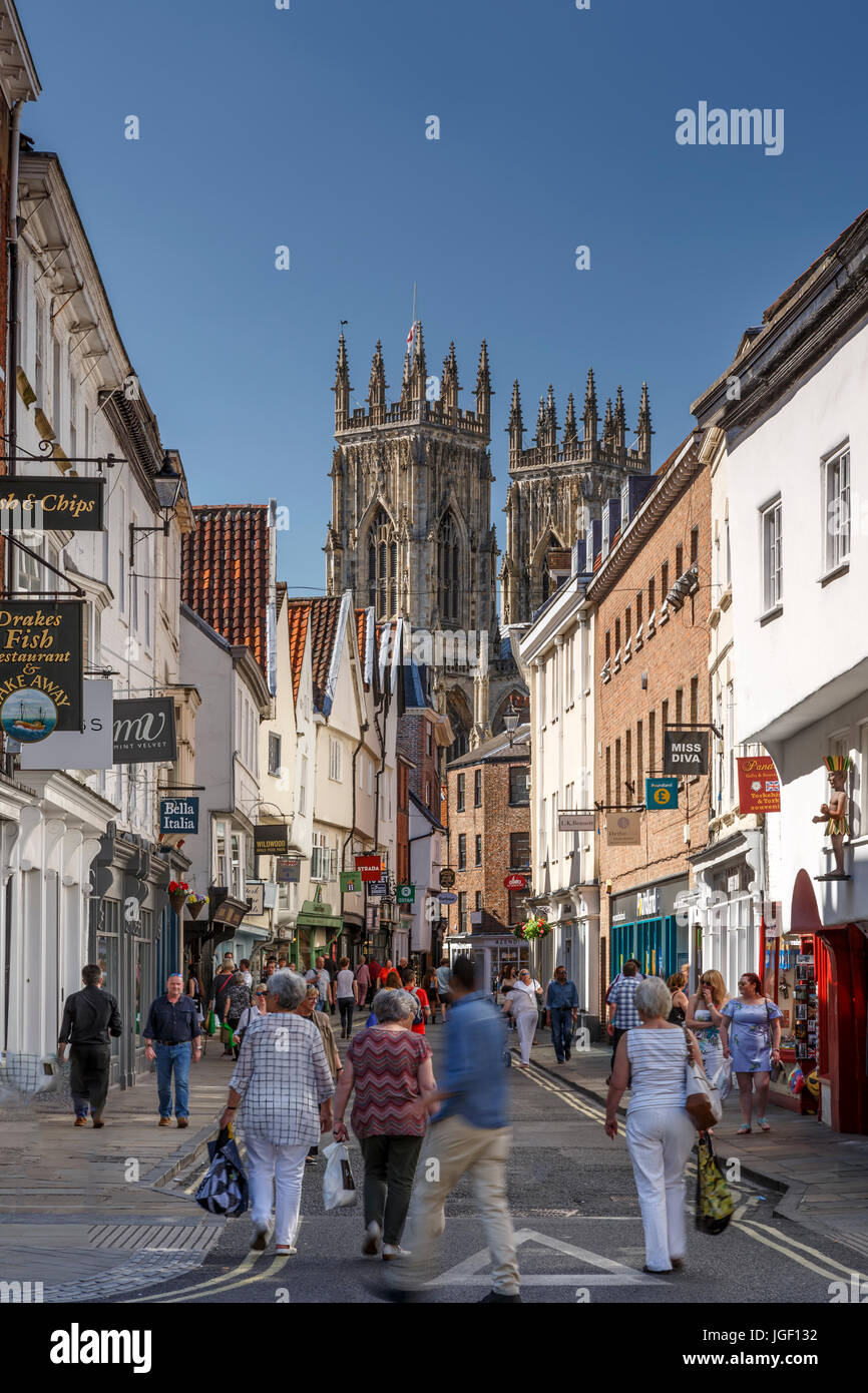 Persone che passeggiano attraverso affollata strada commerciale e le torri della cattedrale di York Minster (la Cattedrale Metropolitical e Chiesa di San Pietro in background, York, Foto Stock