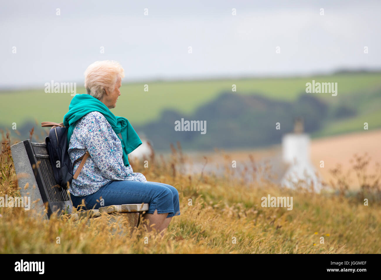 Un lone anziani di sesso femminile solo seduta su una panca in legno lungo il Cornish sentiero costiero tra Polzeath e Daymer Bay in Cornovaglia, Inghilterra cercando Foto Stock
