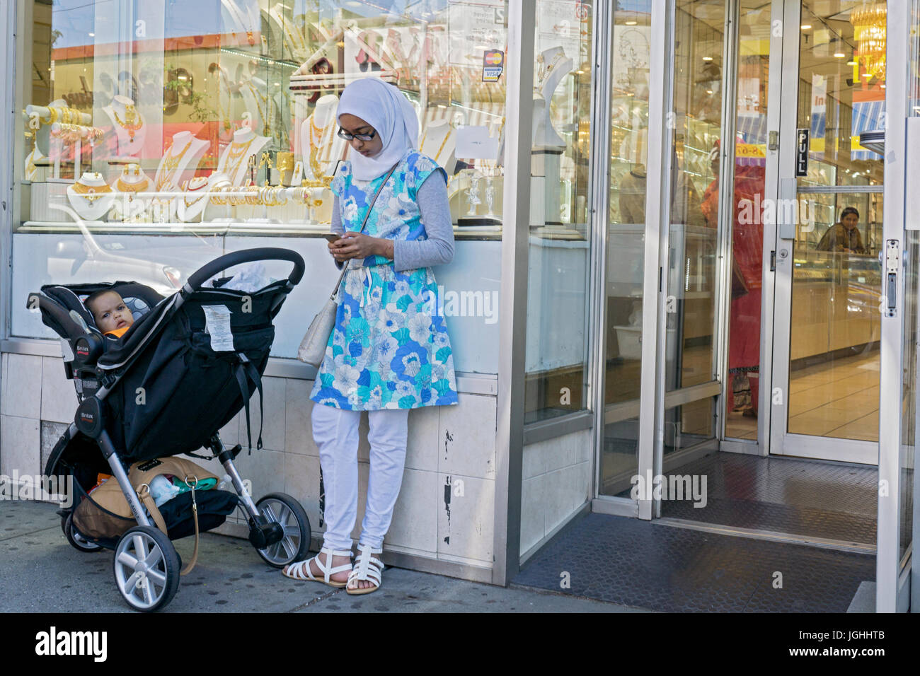 Una giovane donna musulmana guardando il suo telefono cellulare in prossimità di un bambino trasporto nella parte anteriore di un negozio gioielli su 74a san In Jackson Heights, Queens, a New York City Foto Stock