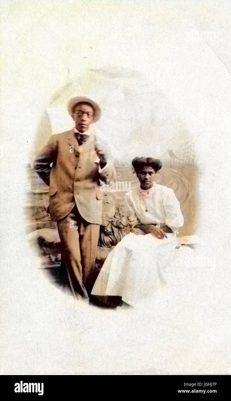Afro-americano di coppia, un uomo e una donna, in posa per un ritratto in studio contro un fondale dipinto, l'uomo che indossa un vestito e un cappello di paglia e in piedi, la donna che indossa un abito bianco e seduto con le braccia incrociate in grembo, entrambi con gravi le espressioni facciali, 1905. Nota: l'immagine è stato colorizzato digitalmente usando un processo moderno. I colori possono non essere periodo-precisa. Foto Stock