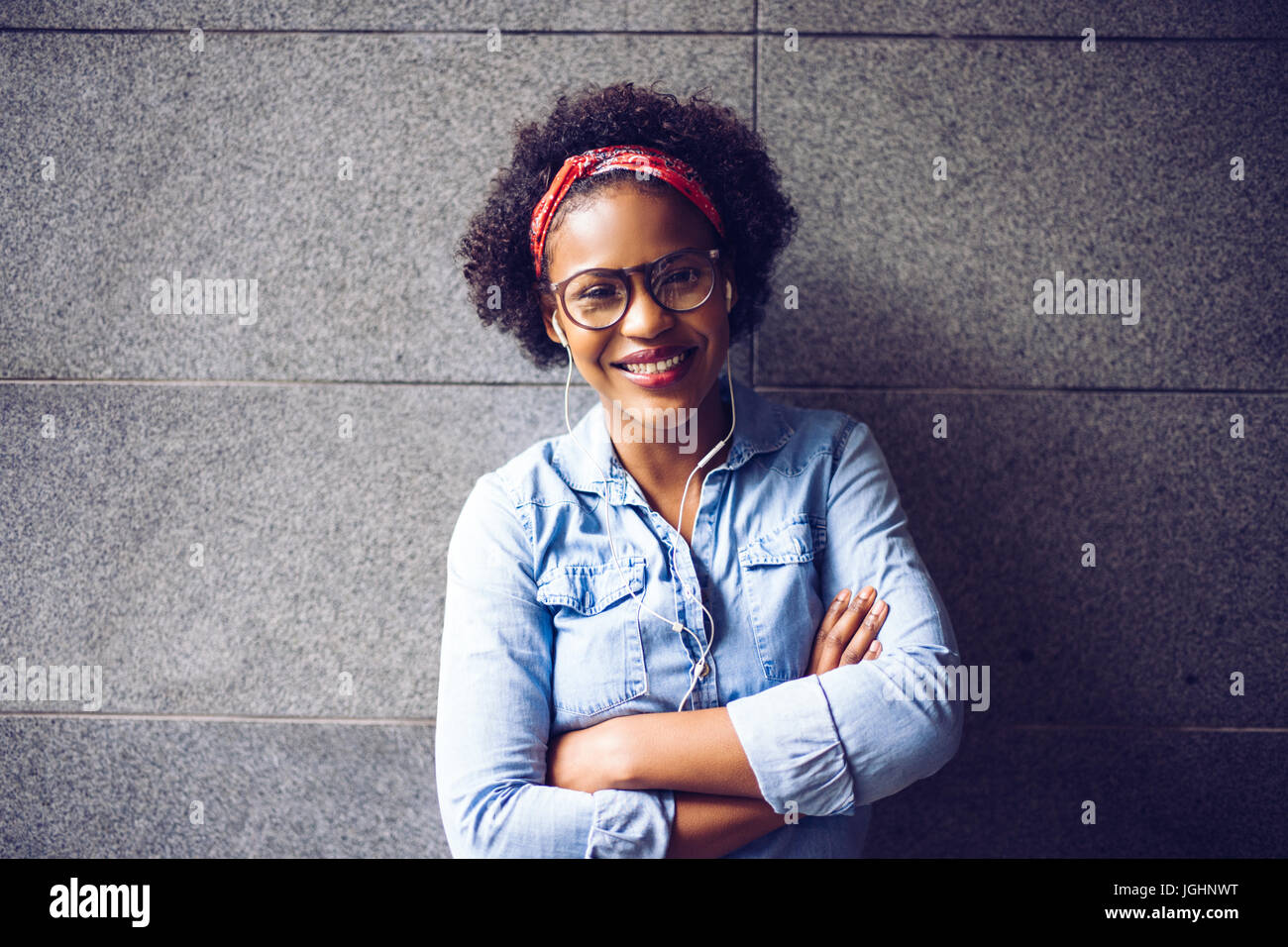 Sorridente giovane donna africana in bicchieri e indossa una bandana in piedi con le braccia incrociate contro una parete ascoltando musica in cuffia Foto Stock