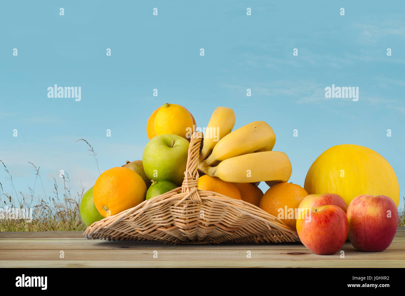 Una selezione di frutta, il riempimento di un fiore in vimini cesto e traboccante su legno tabella planked, con erba selvatica prato e il Cielo di estate blu con sparse Foto Stock