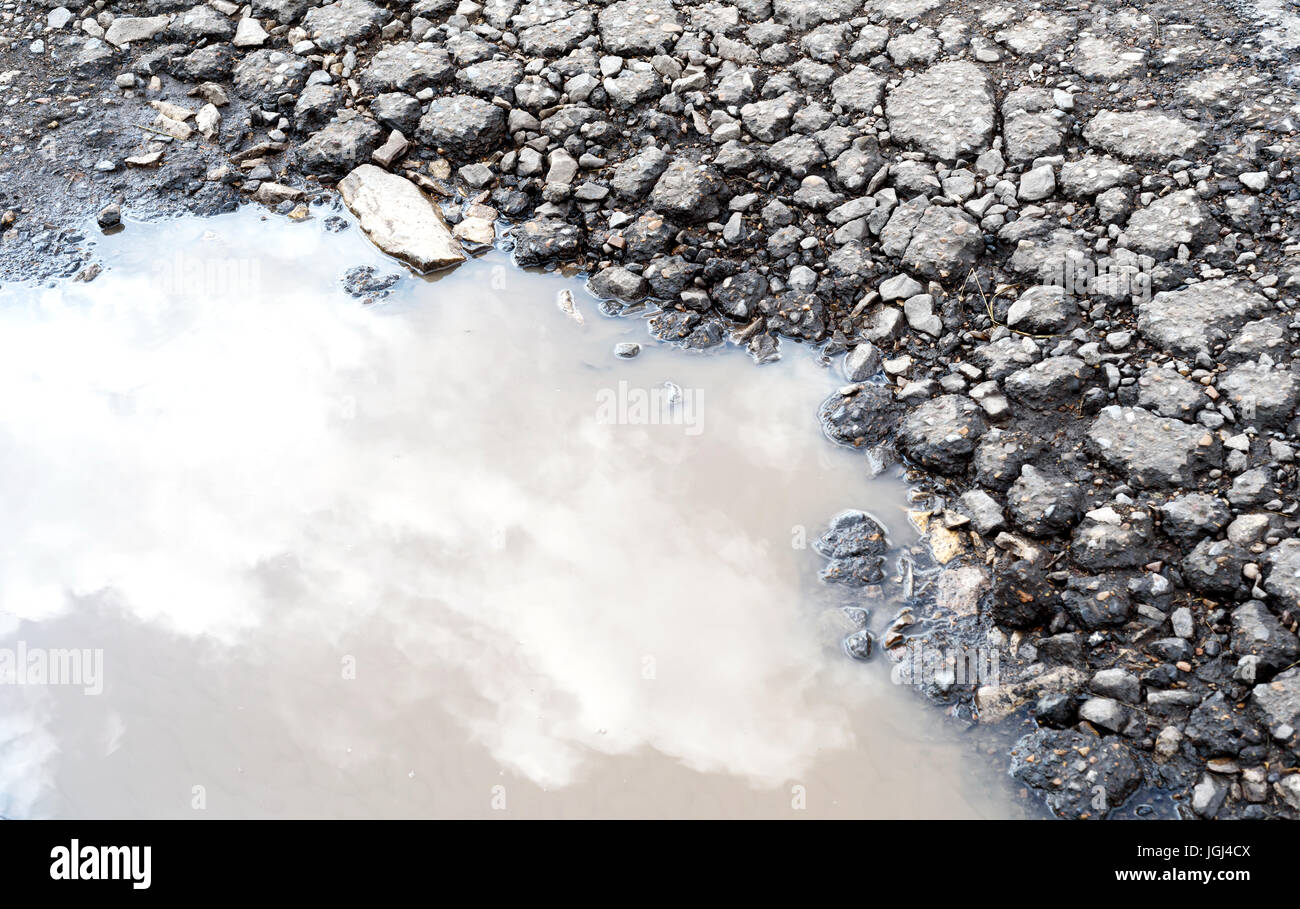 È incrinata e rotture di asfalto buche sulla superficie di una strada fatiscente. Pioggia pozza d'acqua si raccoglie nel foro fessurato che riflette il cielo. Foto Stock