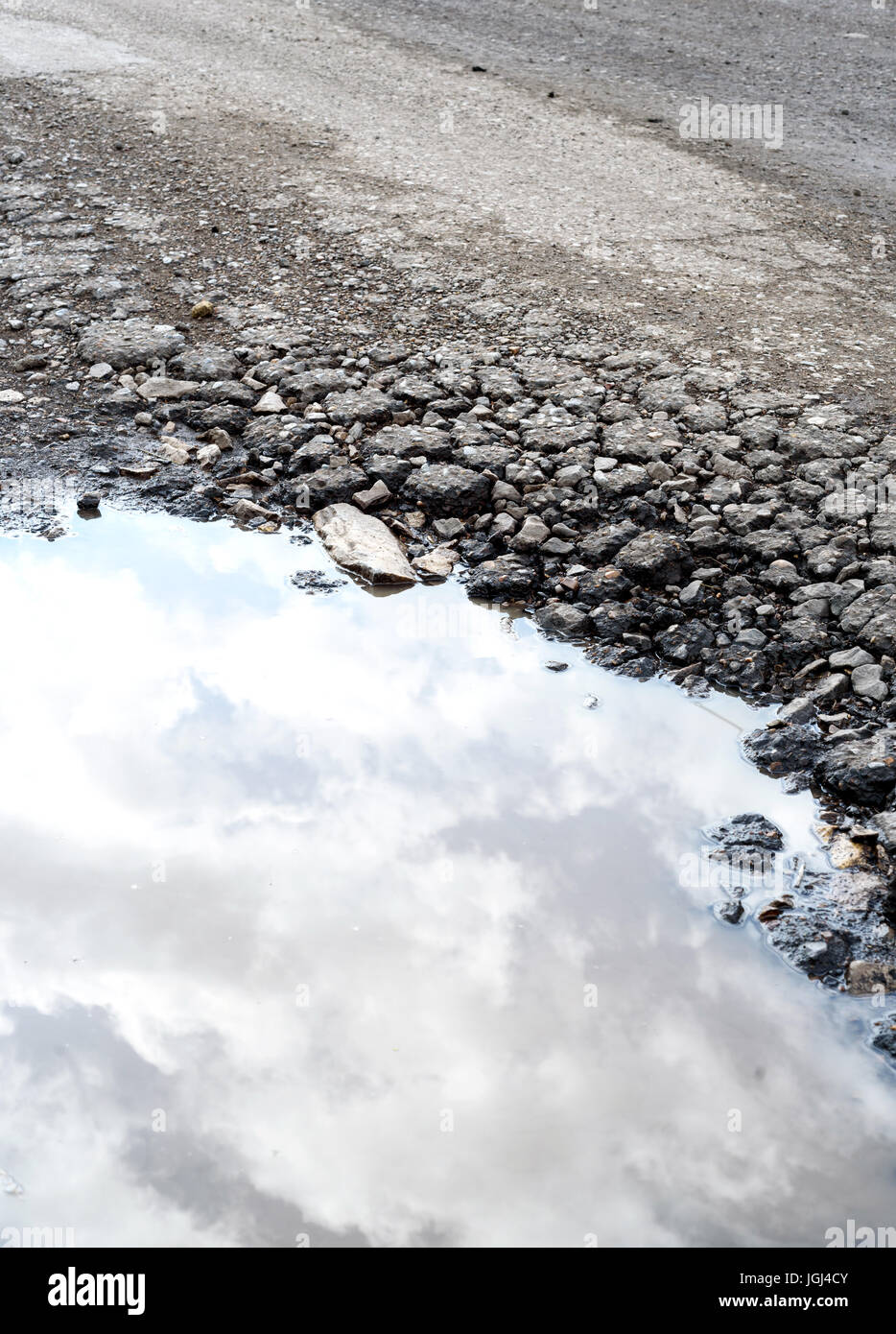 È incrinata e rotture di asfalto buche sulla superficie di una strada fatiscente. Pioggia pozza d'acqua si raccoglie nel foro fessurato che riflette il cielo. Foto Stock