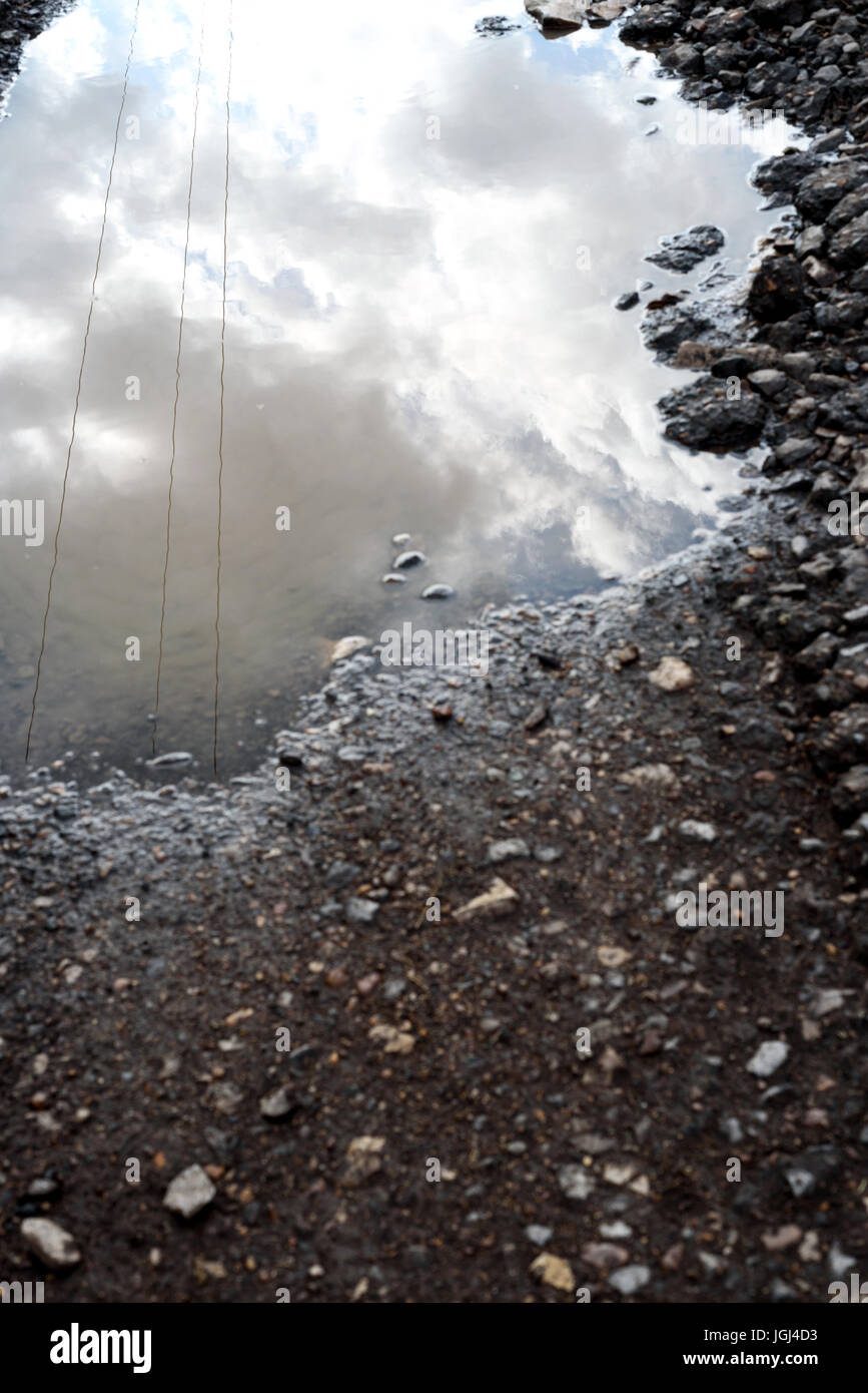 È incrinata e rotture di asfalto buche sulla superficie di una strada fatiscente. Pioggia pozza d'acqua si raccoglie nel foro fessurato che riflette il cielo. Foto Stock