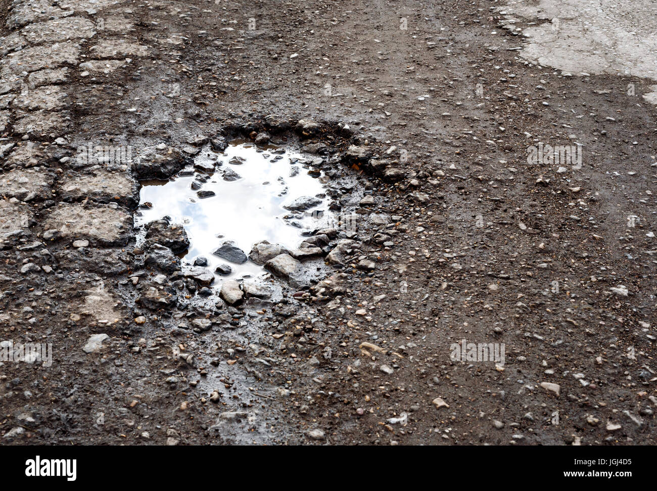 È incrinata e rotture di asfalto buche sulla superficie di una strada fatiscente. Pioggia pozza d'acqua si raccoglie nel foro fessurato che riflette il cielo. Foto Stock