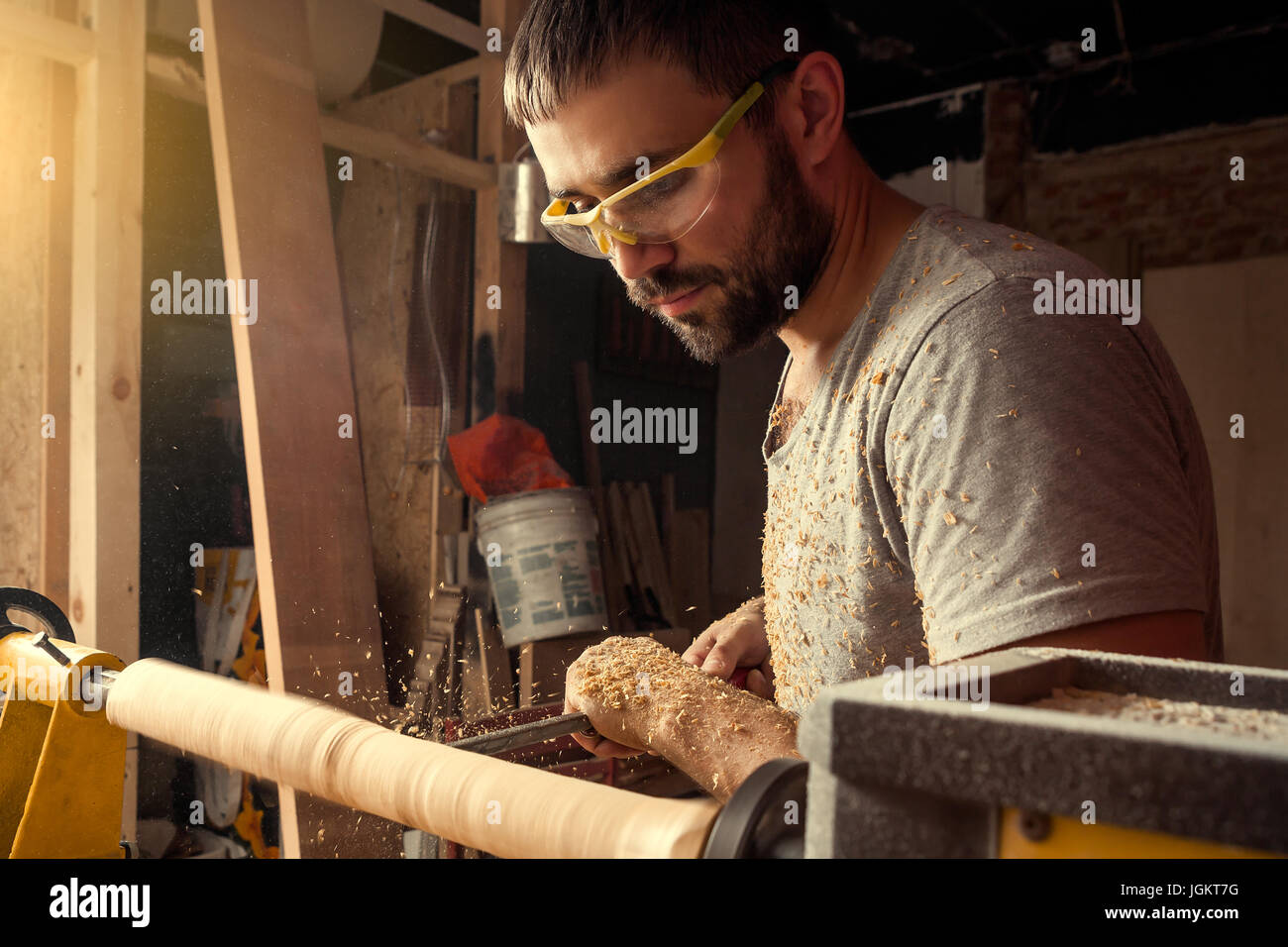 Un giovane uomo builder indossare gli occhiali di protezione con la barba e grigio di una t-shirt gestisce l'albero su un tornio in officina, sullo sfondo di un sacco di woo Foto Stock