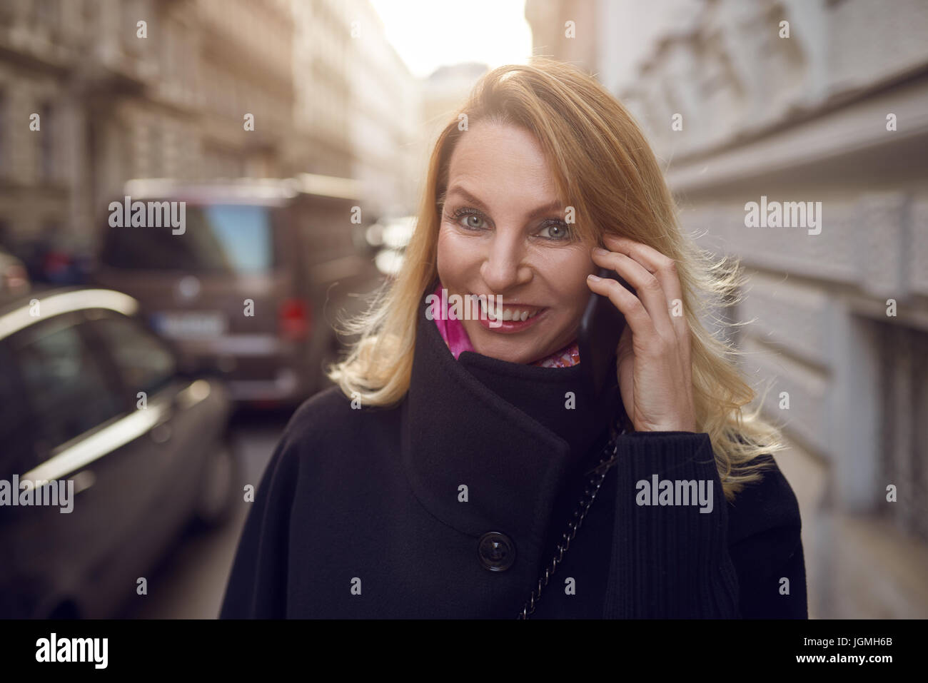 Piuttosto vivace donna chattare su un telefono mobile come lei cammina giù per una strada urbana sorridente felicemente con la videocamera Foto Stock