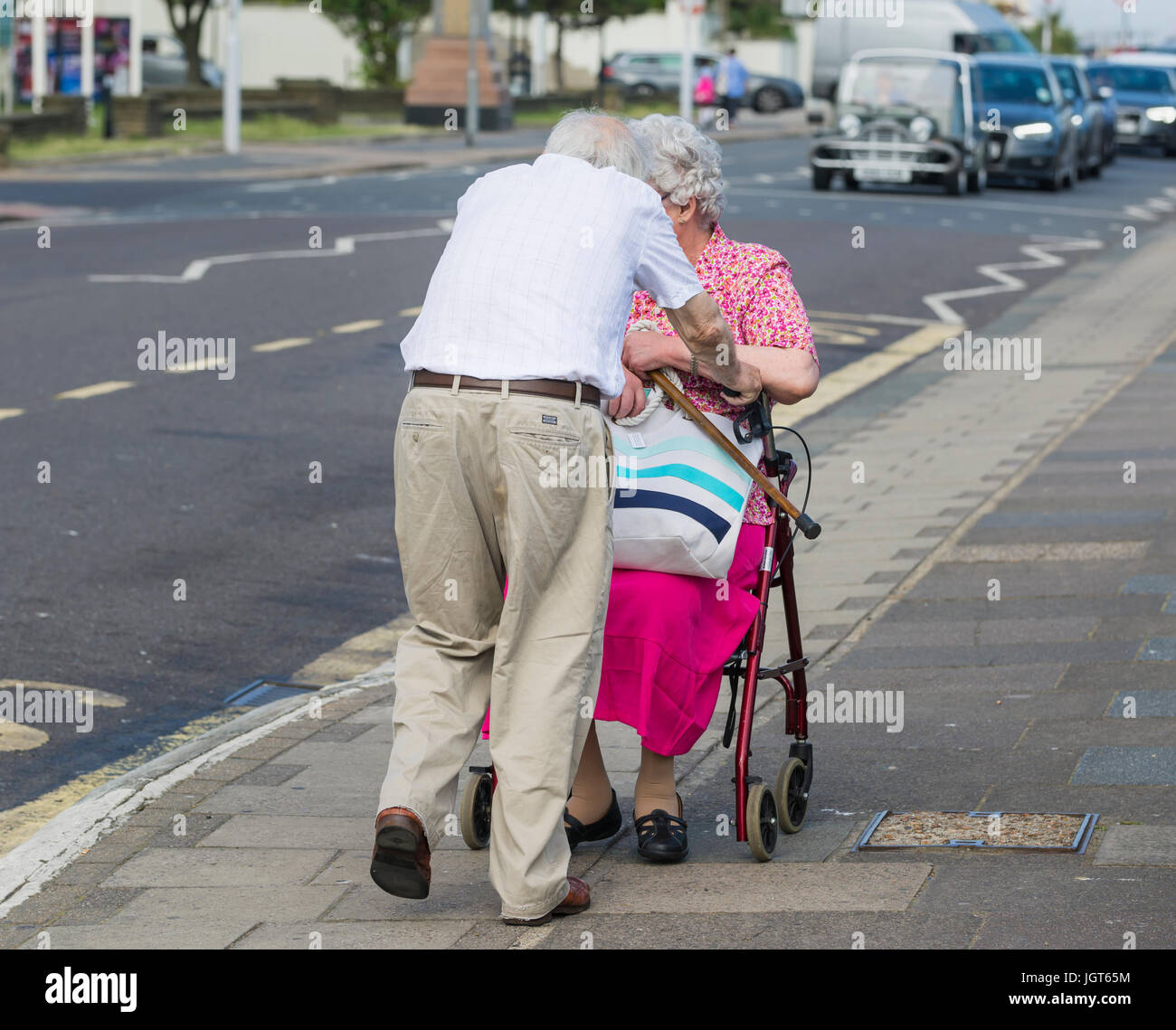 Coppia di anziani che si aiutano reciprocamente a piedi lungo, con una sola seduta su ruote telaio a piedi. Foto Stock