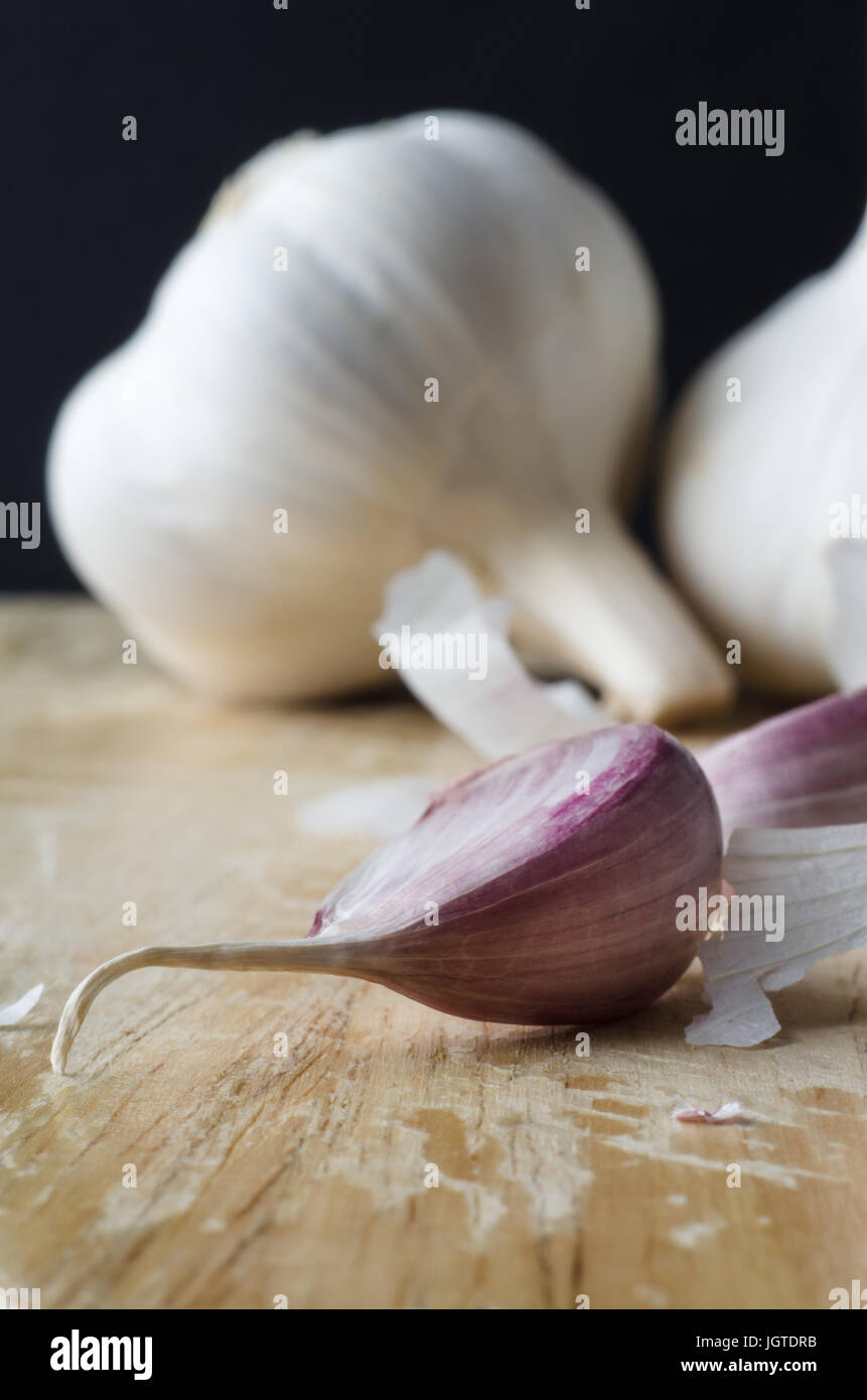 Due interi bulbi di aglio in background, con chiodi di garofano rosa in primo piano su un tagliere di legno circondato da sparsi frammenti di shedded pape Foto Stock