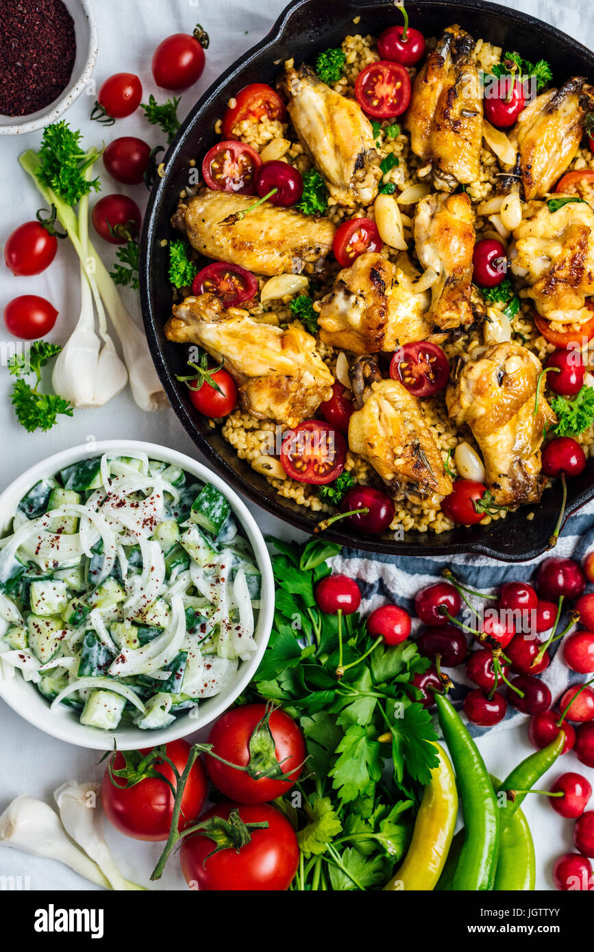 Ali di pollo e Bulgur pilaf cucinato in una padella, guarnita con ciliegie, pomodorini e erbe fotografata da vista dall'alto. Foto Stock