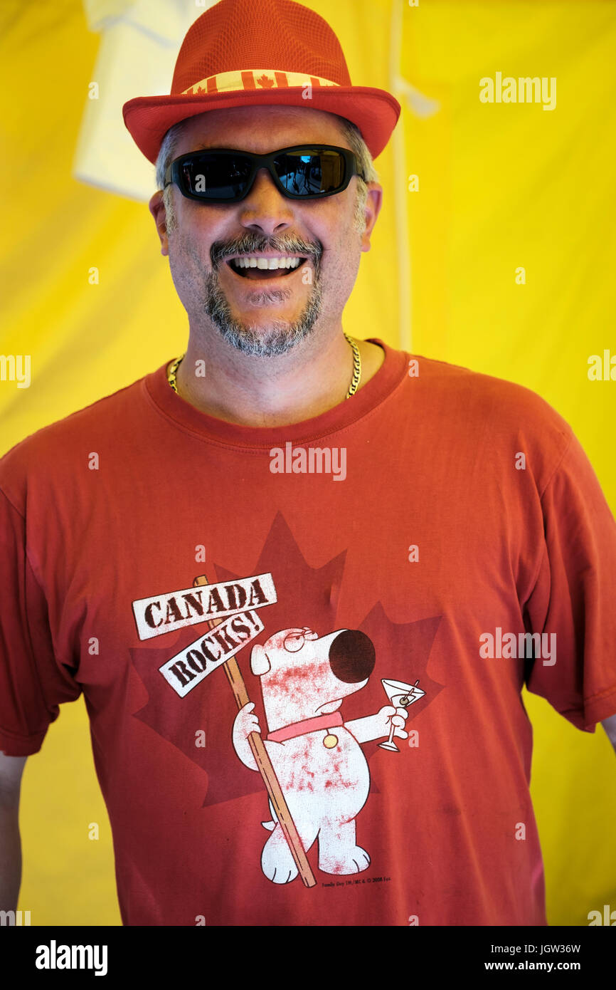 Uomo di mezza età con occhiali da sole, canadese themed hat, t-shirt guardando la telecamera, sorridente alla fotocamera, celebrando il Canada per il 150° compleanno. Foto Stock