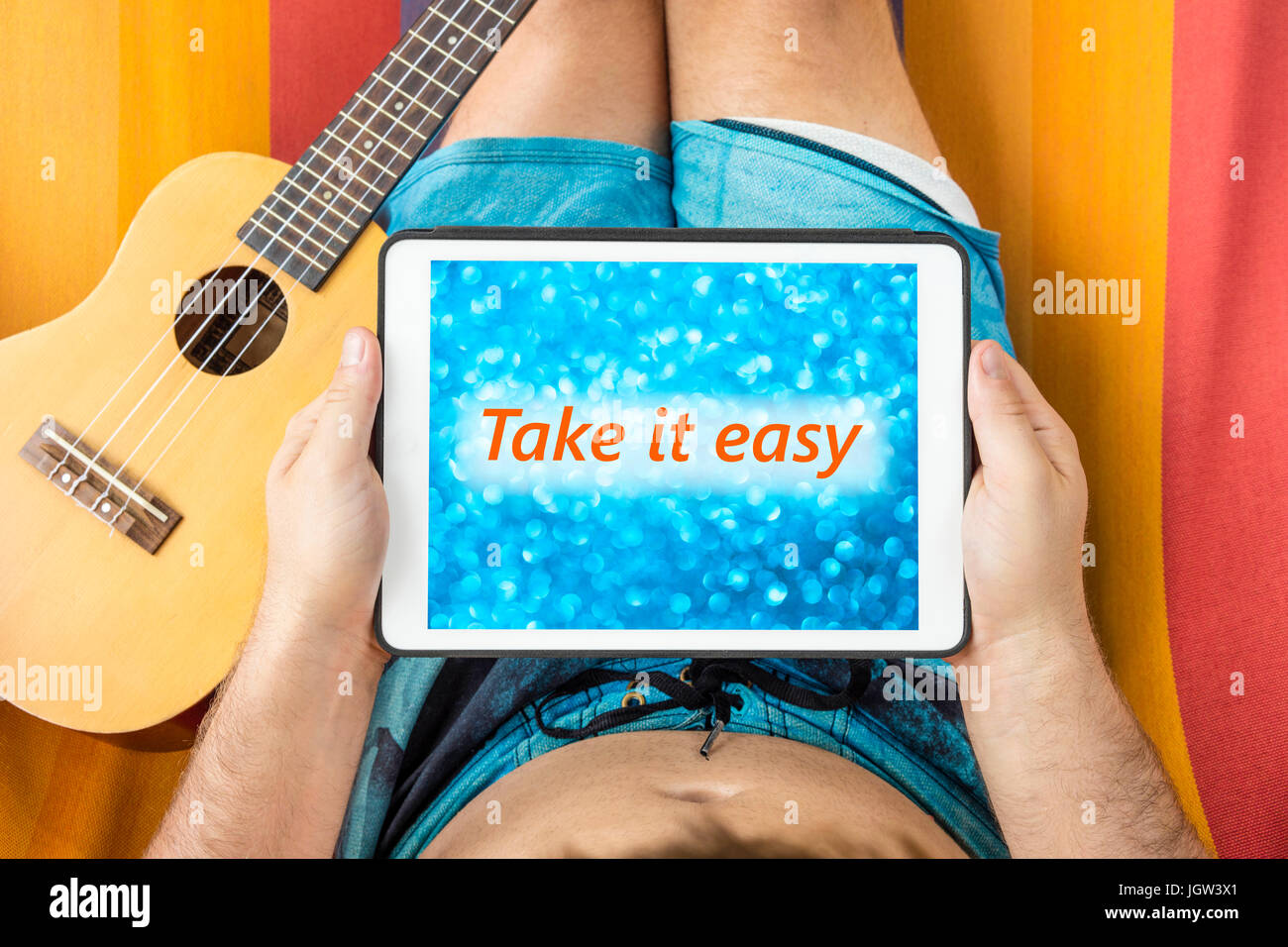Giovane uomo disteso su una amaca con dispositivo tablet guardando sfocato sfondo blu con la frase "Take it easy' scritto su di esso. Foto Stock