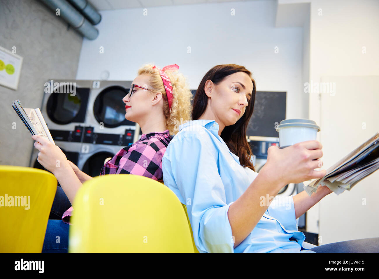 Due donne leggendo quotidiani di schiena nella lavanderia a gettoni Foto Stock