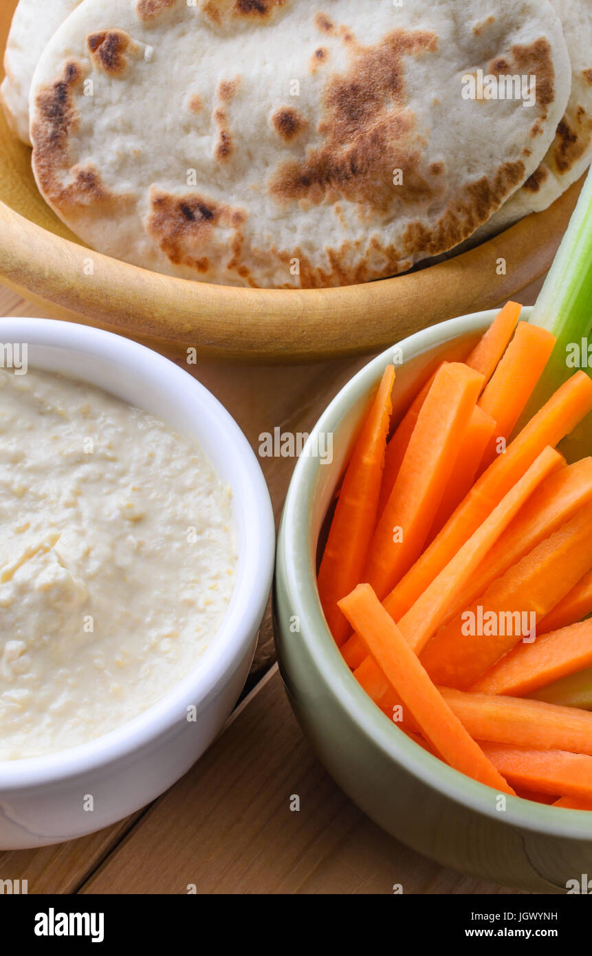 Close up di ciotole contenenti hummus dip; mini pane pitta e la carota e il sedano crudites su un asse di legno tabella. Preso dal Diner's prospettiva Foto Stock