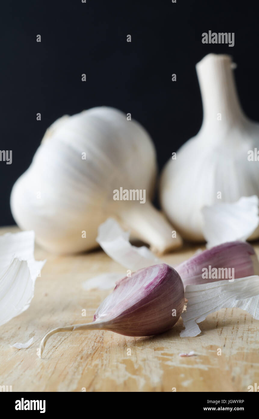 Due interi bulbi di aglio in background, con chiodi di garofano rosa in primo piano su un tagliere di legno circondato da sparsi frammenti di shedded pape Foto Stock
