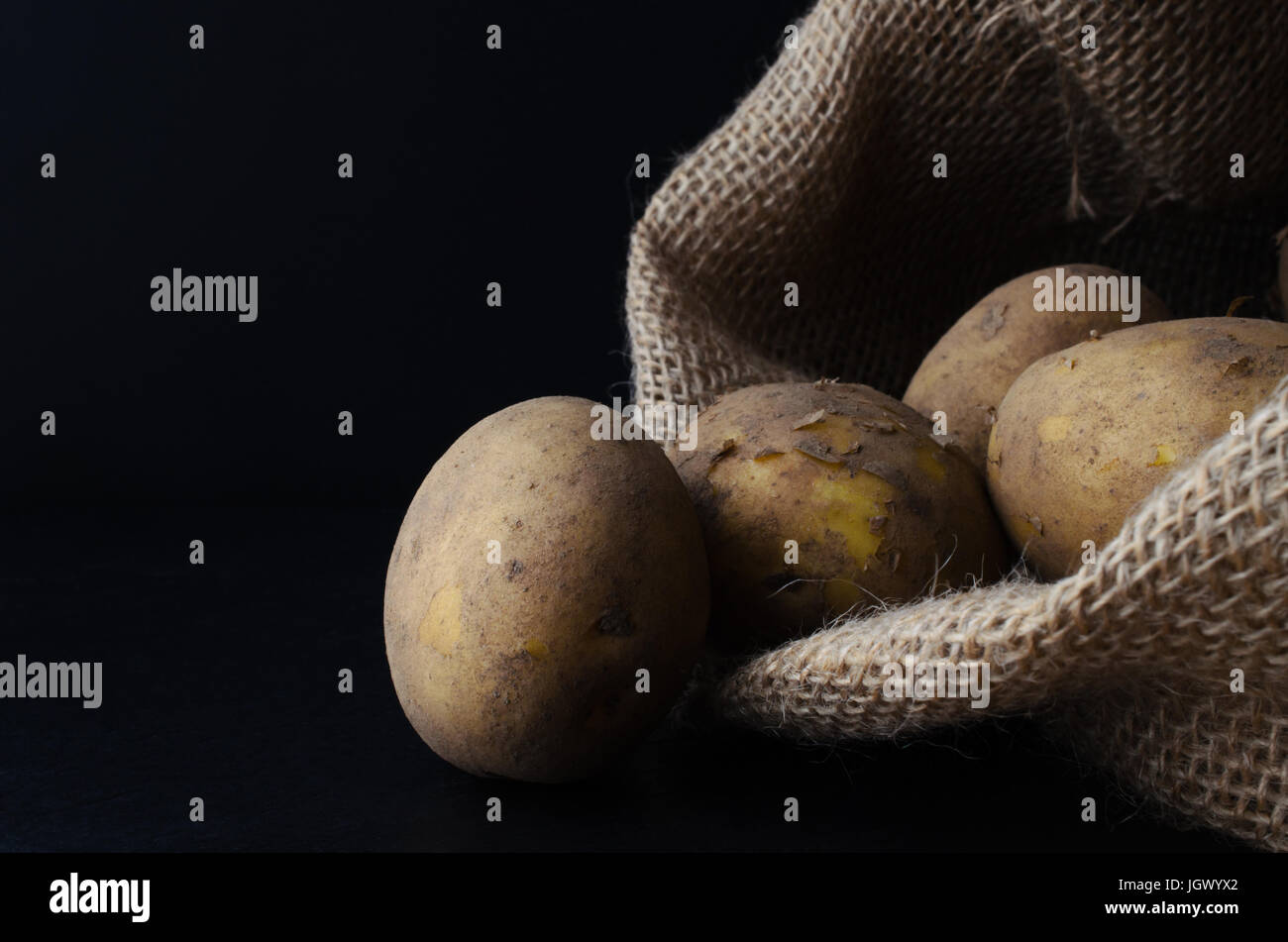Materie, non lavate e con la buccia di patate, la fuoriuscita del sacco di Hesse al nero ardesia superficie. Moody illuminazione, sfondo nero. Foto Stock