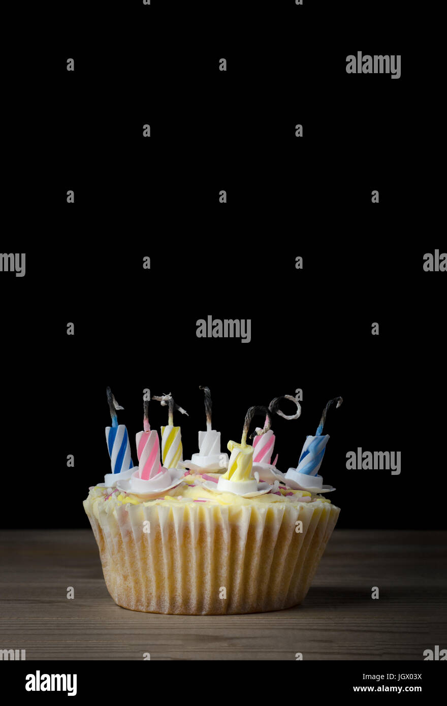 Un compleanno cupcake in un semplice caso di carta con sette candele a strisce che hanno incendiato e stato bruciato. Posto su una tavola di legno con nero b Foto Stock