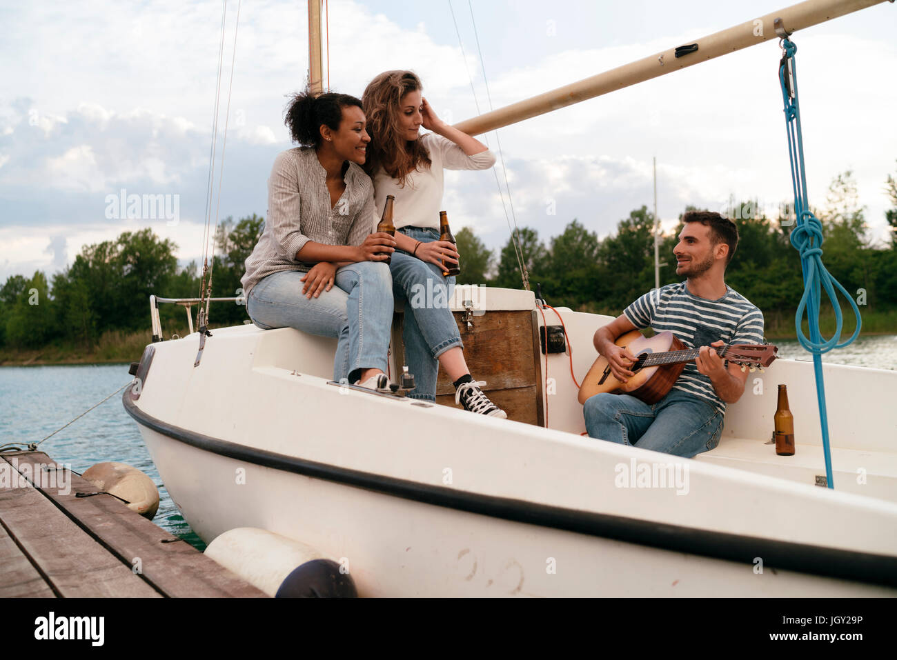 Tre amici rilassante ormeggiata sulla barca a vela, bere birra, l uomo a suonare la chitarra Foto Stock