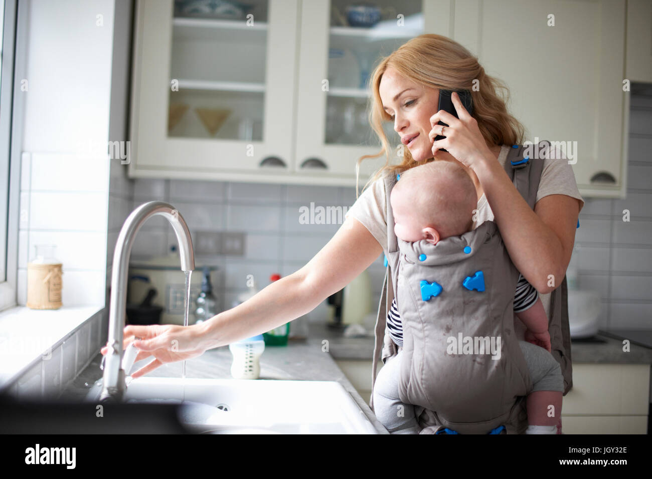 Donna che mantiene baby boy in imbracatura, utilizza lo smartphone, ruotando il rubinetto per fare il lavaggio fino Foto Stock