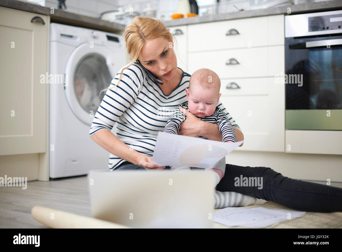 Donna che mantiene il bambino figlio di armi, mentre guardando attraverso il lavoro sul pavimento della cucina, parlando su smartphone e guardando il computer portatile Foto Stock
