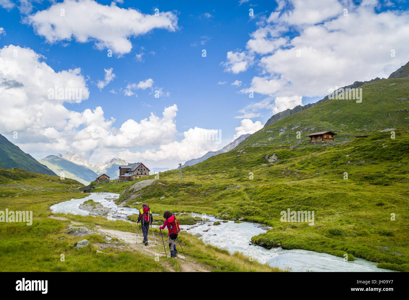 Un paio di escursionisti in direzione di un rifugio di montagna lungo un flusso nelle Alpi sotto un azzurro cielo nuvoloso. Foto Stock