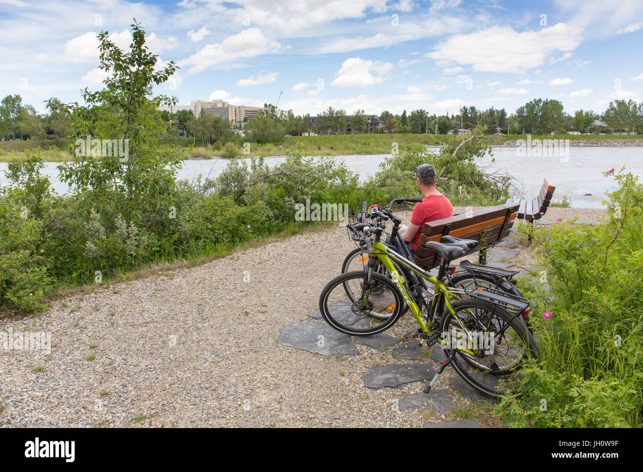 Un uomo è seduto sulla panchina di Edworthy Park con due bici accanto a lui lungo il Fiume Bow a Calgary, Alberta, Canada Foto Stock