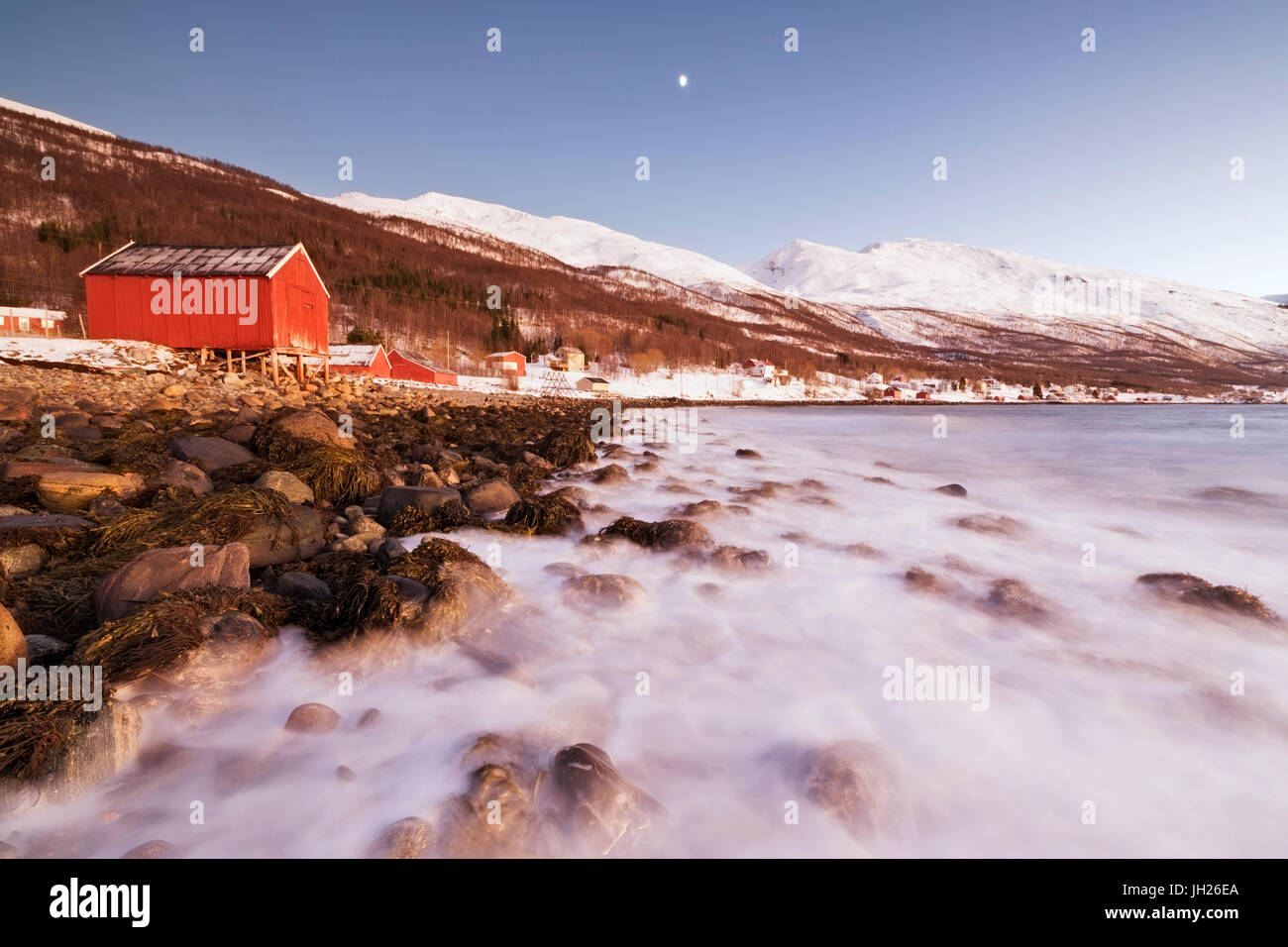 Onde del mare freddo che si infrangono sulle rocce e tipiche baite in legno chiamato Rorbu, Djupvik, Alpi Lyngen, Troms, Norvegia e Scandinavia Foto Stock