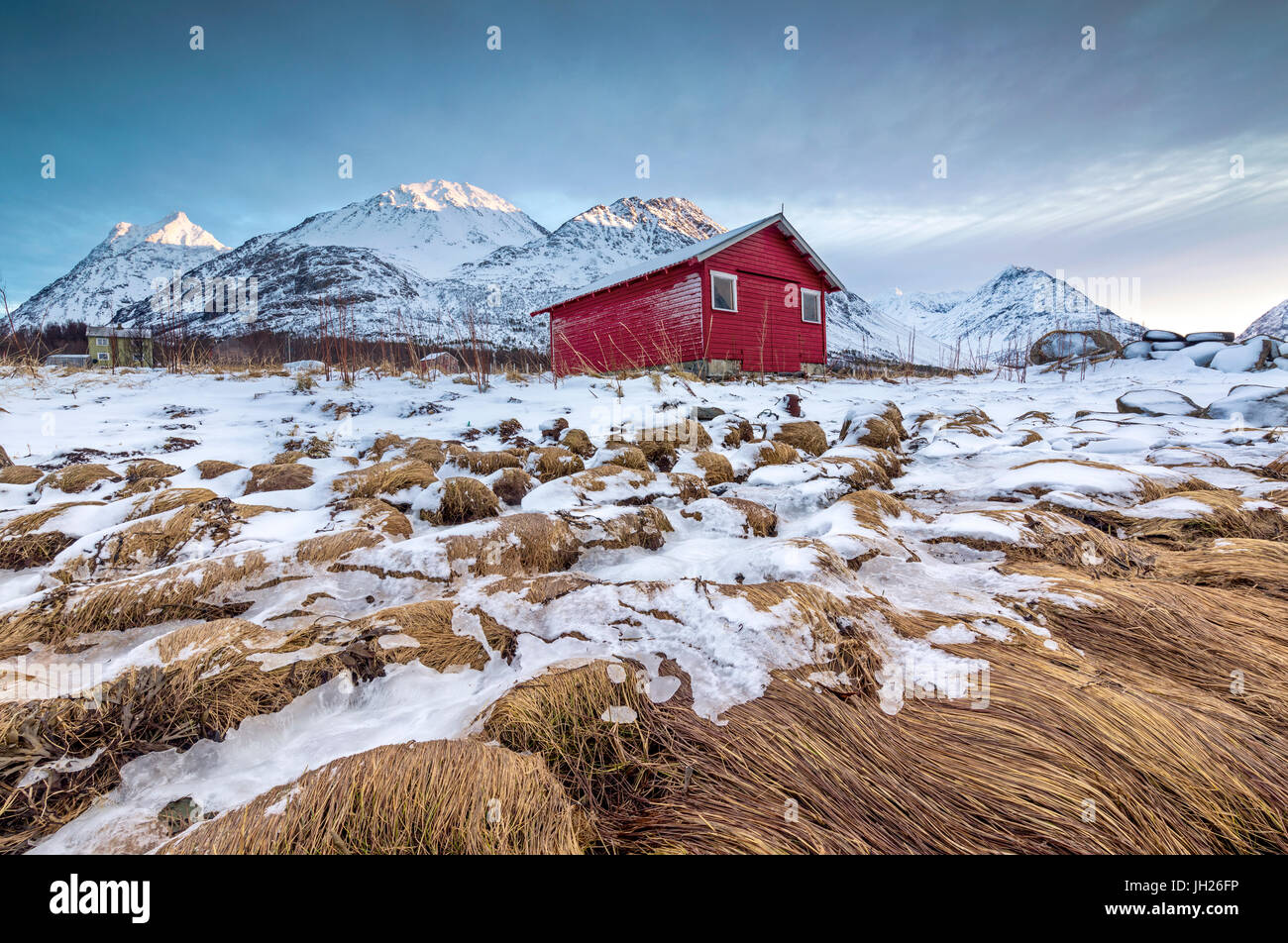 Capanna in legno incorniciata da rocce ricoperte di erba e il ghiaccio con cime innevate sullo sfondo, Svensby, Alpi Lyngen, Troms, Norvegia Foto Stock