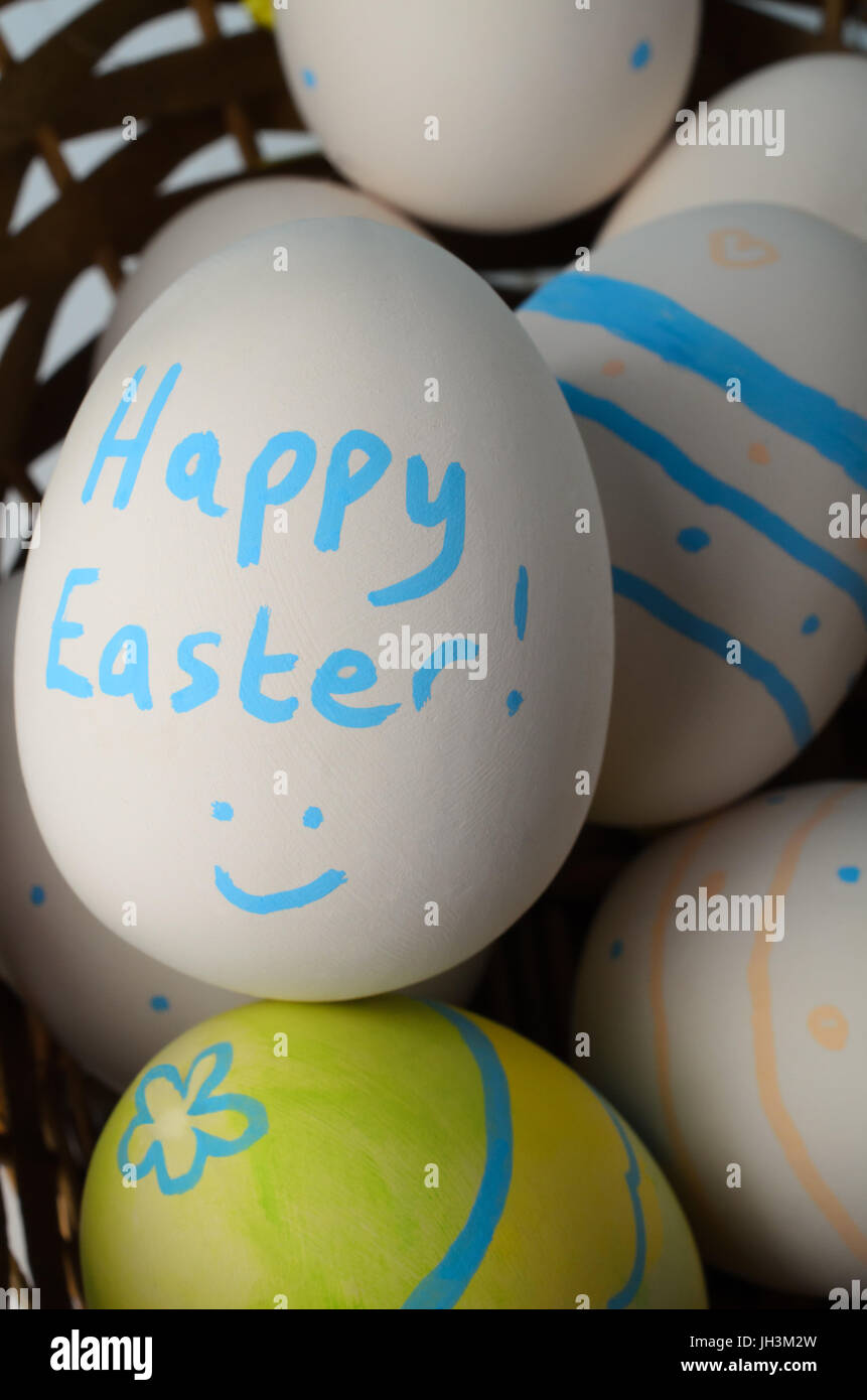 Close up, overhead shot di più bianco crema di uova di ceramica in un biscotto di vimini, decorati con motivi, un volto sorridente e le parole 'Buona Pasqua Foto Stock