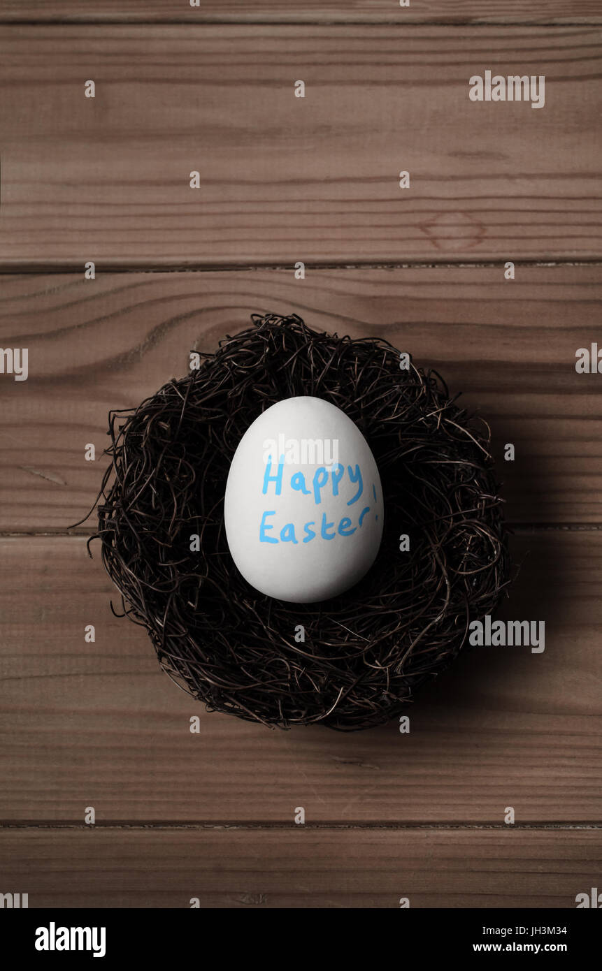 Concetto di pasqua. Tettuccio di colpo di un falso uovo bianco in un nido sul tavolato di legno. Il messaggio 'Felice Pasqua' è scritto sull'uovo in blu. Tonica per un Foto Stock