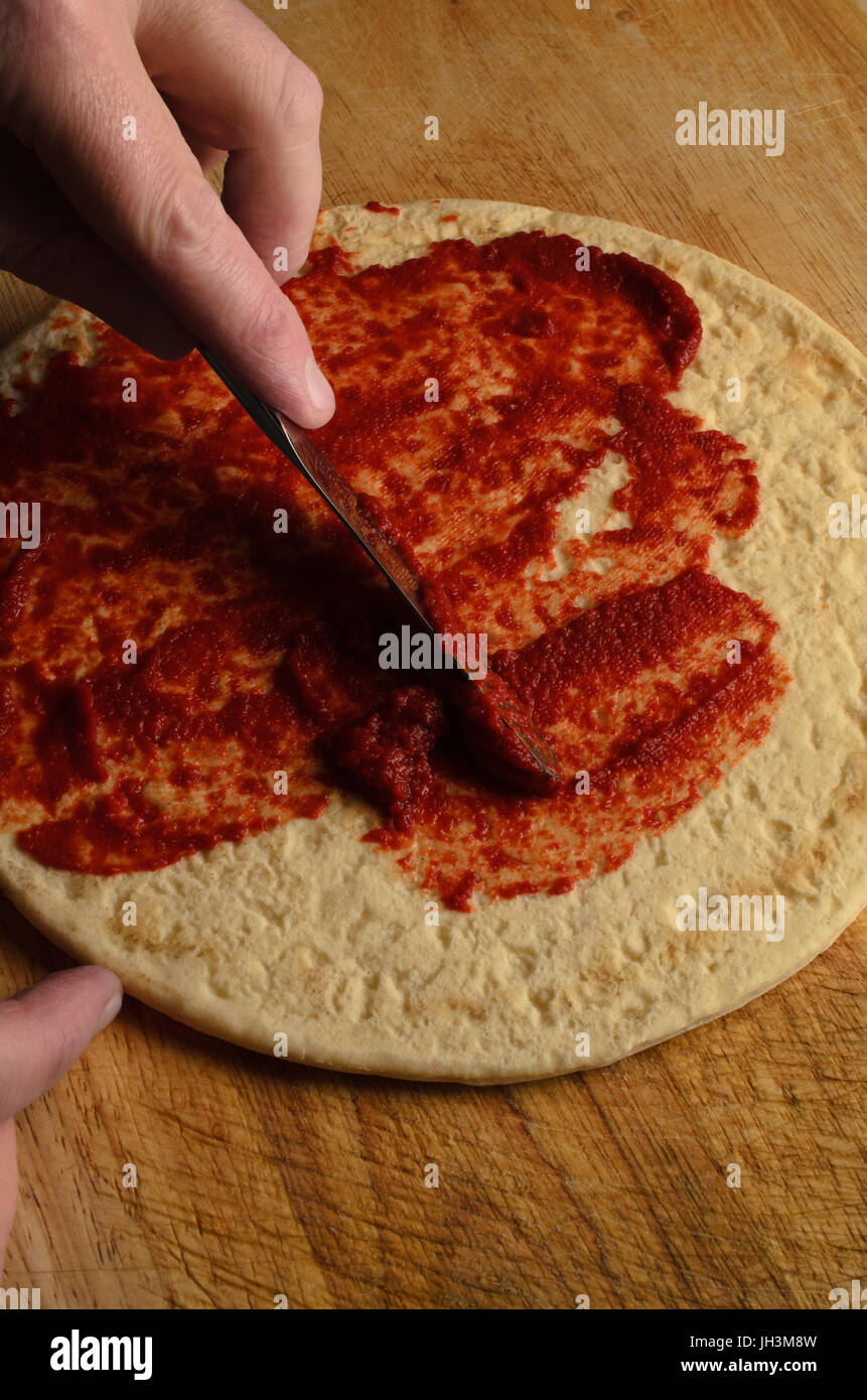 Un maschio di spargimento a mano la salsa di pomodoro sulla base per pizza con il coltello su un vecchio, graffiato e indossato tagliere di legno. Luce oscura. Foto Stock