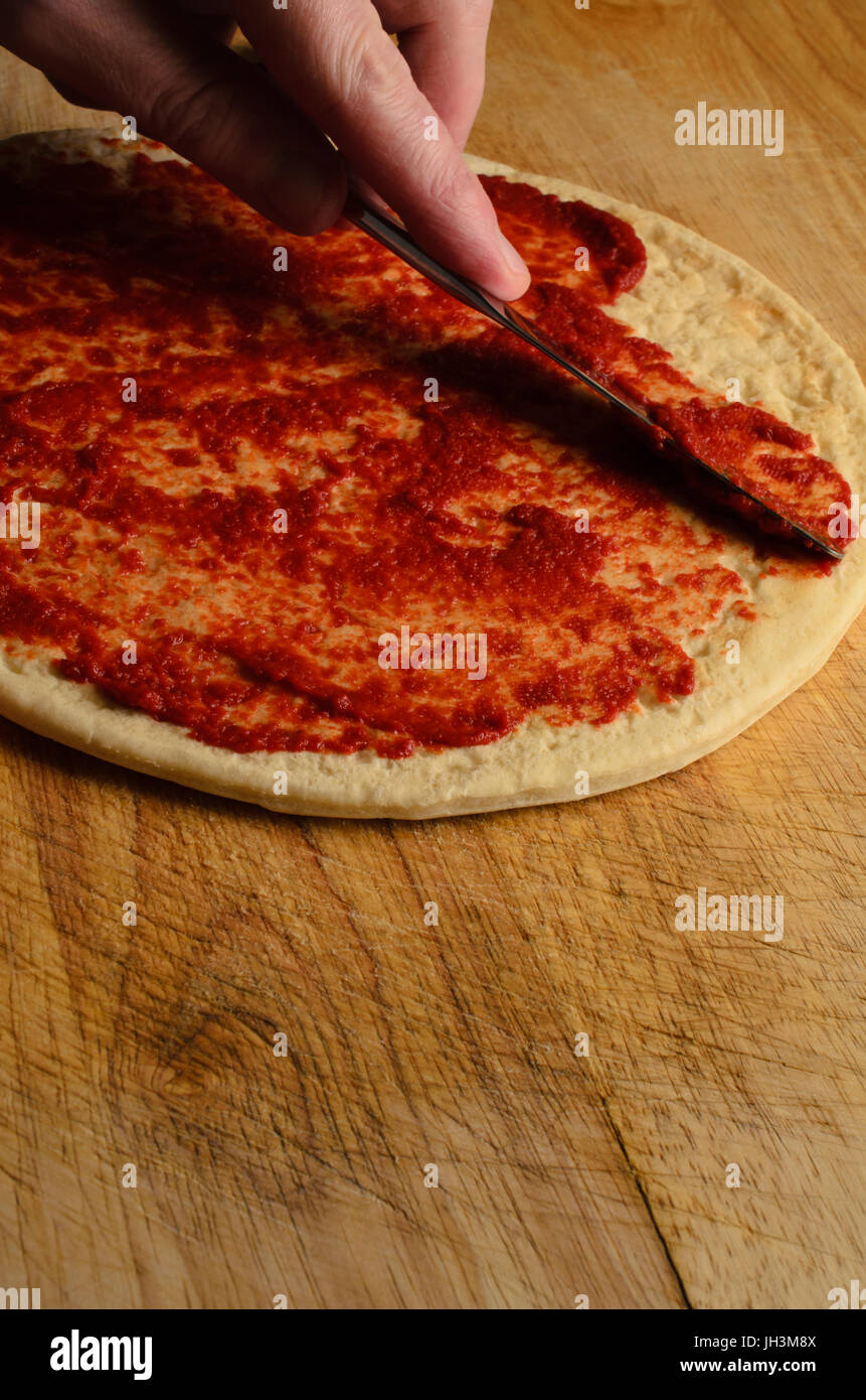 Un maschio di spargimento a mano la salsa di pomodoro sulla base per pizza con il coltello su un vecchio, graffiato e indossato tagliere di legno. Luce oscura. Foto Stock