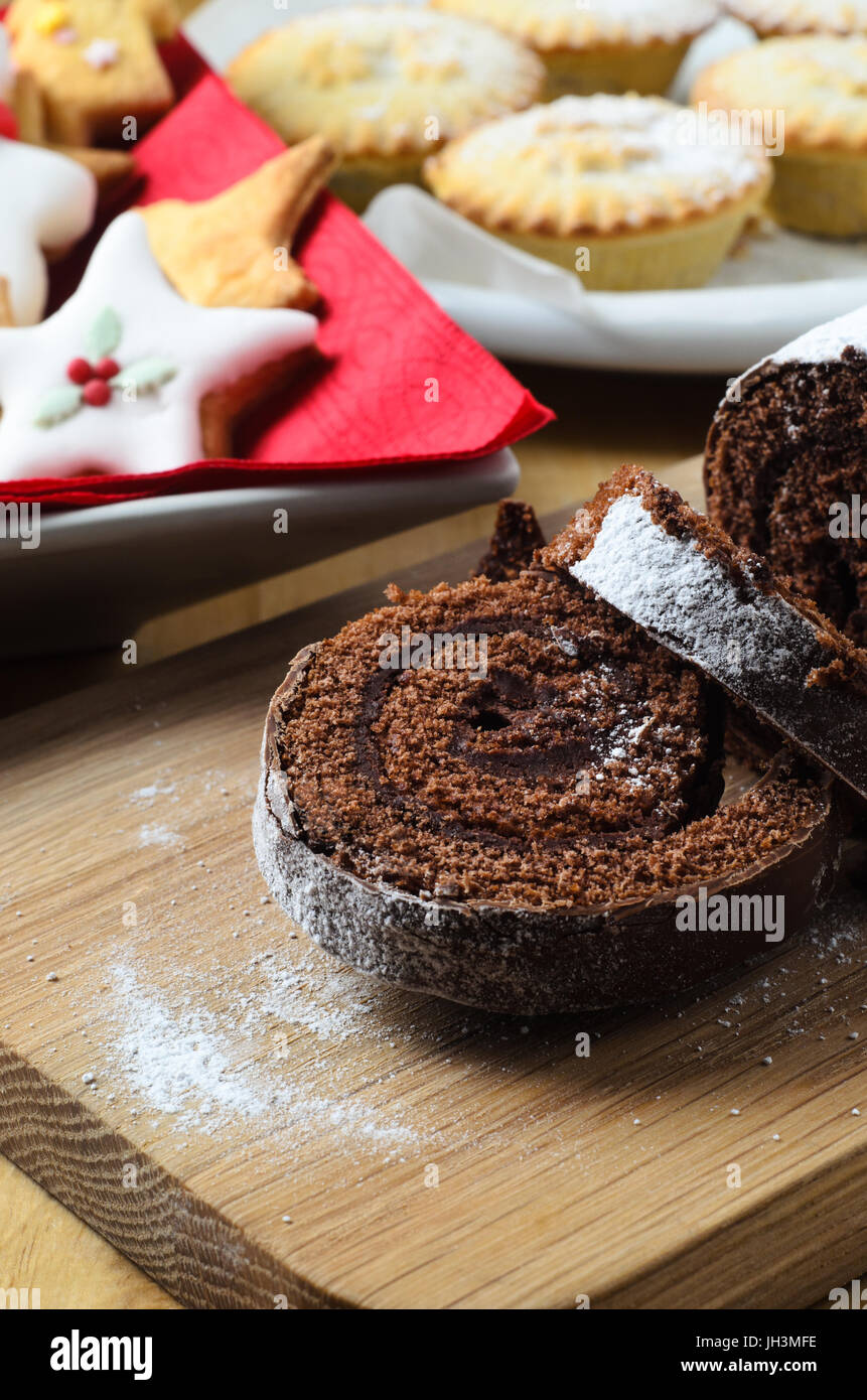 Natale al cioccolato Yule Log su pannello di legno decorato con biscotti e torte di carne macinata in background. Foto Stock