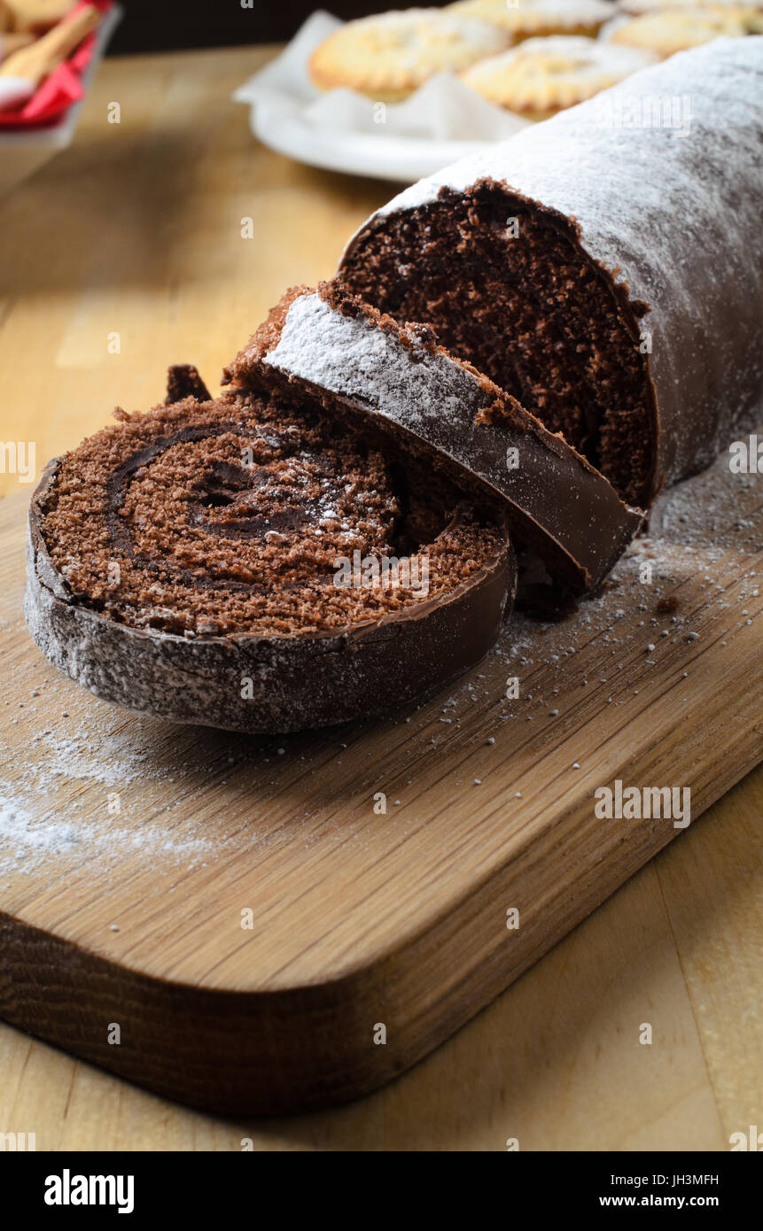 I dolci del Natale tavola con fette di cioccolato Yule Log spolverato con zucchero a velo in primo piano. Tritare torte e biscotti in background. Foto Stock