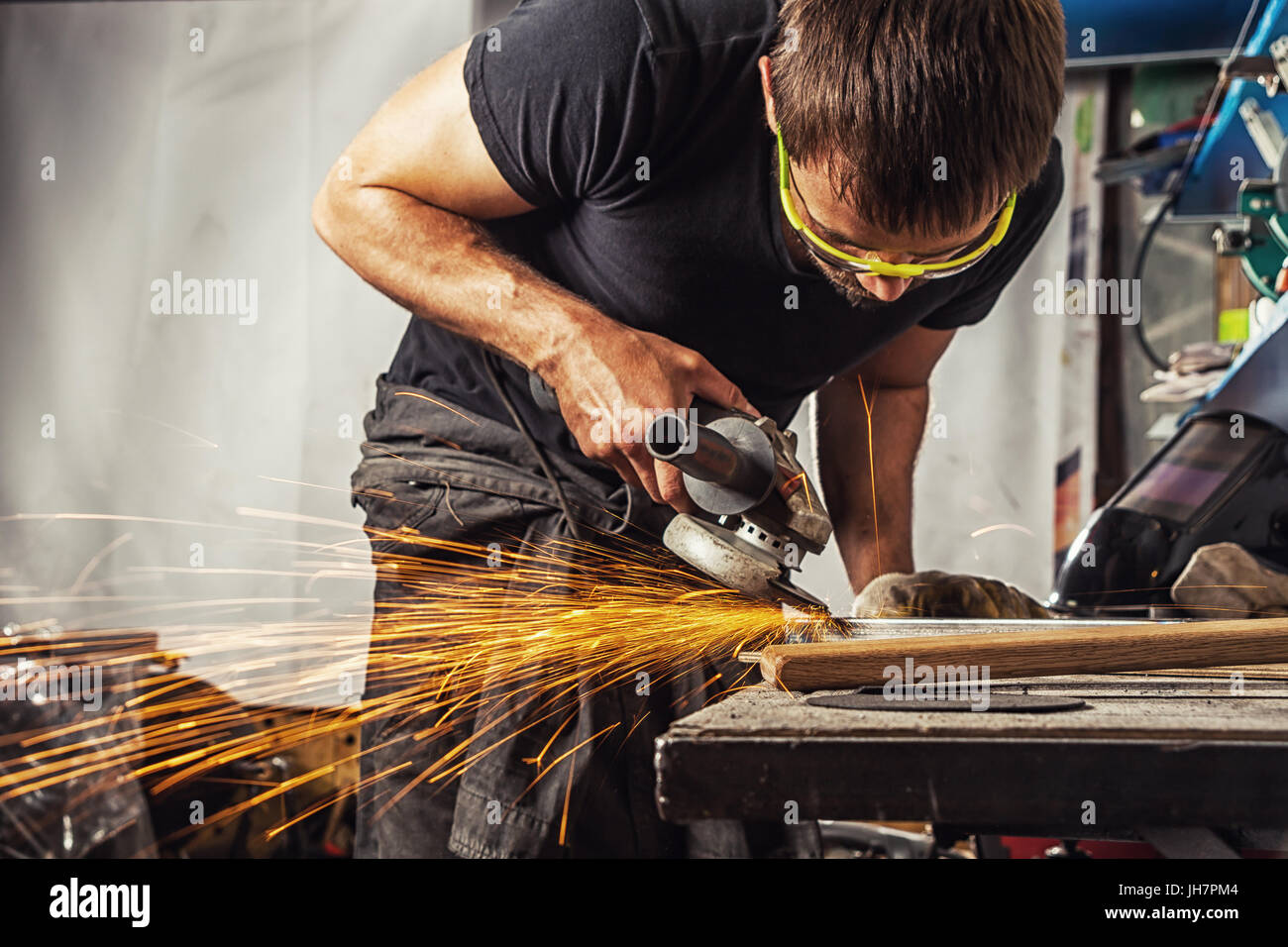 Un giovane uomo è una bruna saldatore indossare una t-shirt nera e occhiali di sicurezza la molatura di metallo con una smerigliatrice angolare in officina, scintille volanti nell' Foto Stock