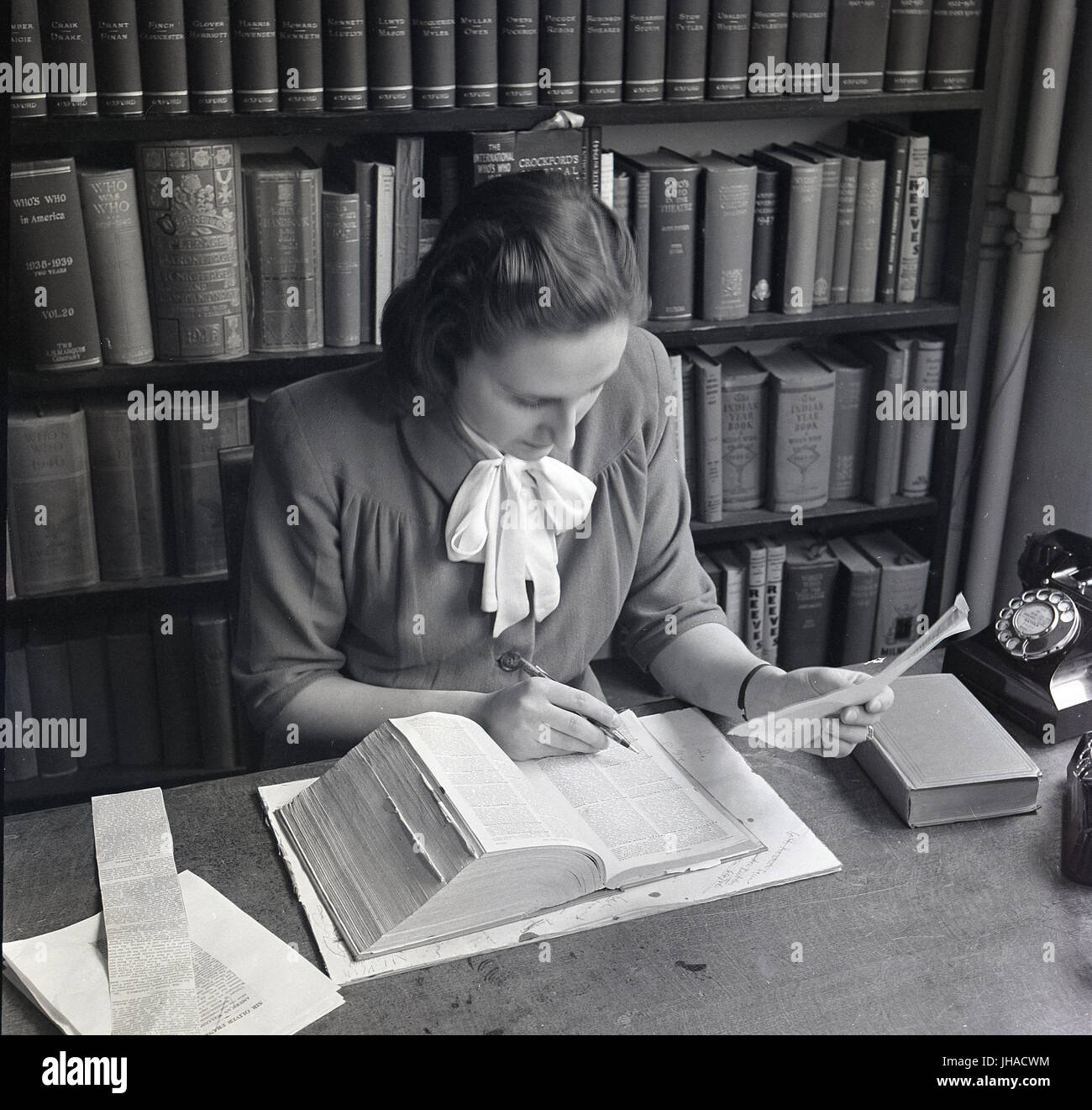1948, storica, giovane donna con penna, libro di testo e telefono a una scrivania che lavora in un ufficio pieno di libri, che controlla i dettagli sulla stampa, British Council, Londra, Inghilterra, Regno Unito. Foto Stock