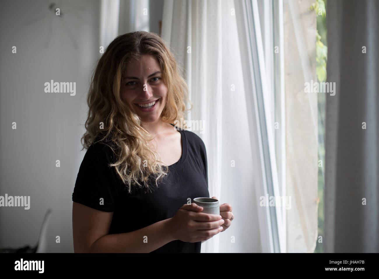 Giovani, Bionda, bella donna caucasica sorseggiando una bevanda calda vicino alla finestra naturale nella luce del mattino.catturato interni durante l'estate su una Canon 5D Foto Stock