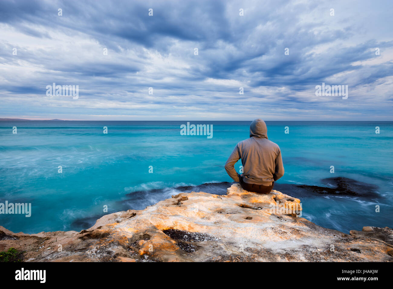 Un uomo si siede sul bordo di un robusto roccia calcarea e si affaccia su un oceano tempestoso vista in Sud Australia. Foto Stock