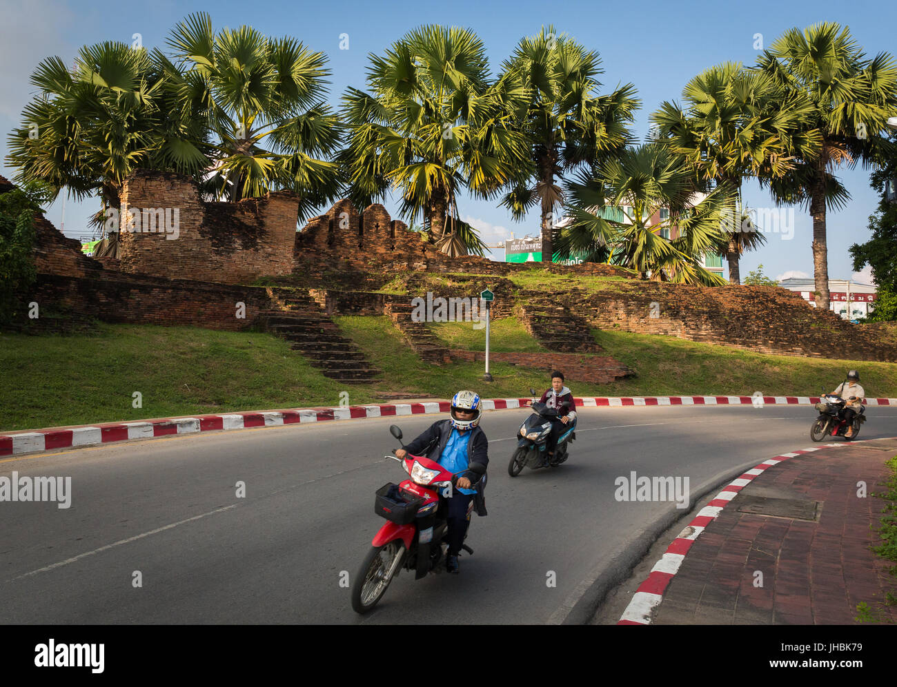 Tre persone andare in motocicletta intorno ad un angolo a Chiang Mai, Thailandia, con alberi di palma e il muro della città vecchia in background. Foto Stock