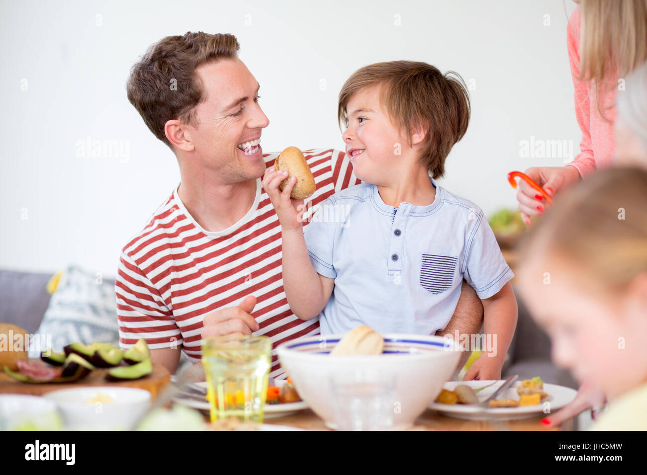 Little Boy è seduta sul suo papà in giro al tavolo da pranzo in casa loro a ora di cena. Egli sta cercando di alimentare il suo padre una pagnotta di pane. Foto Stock