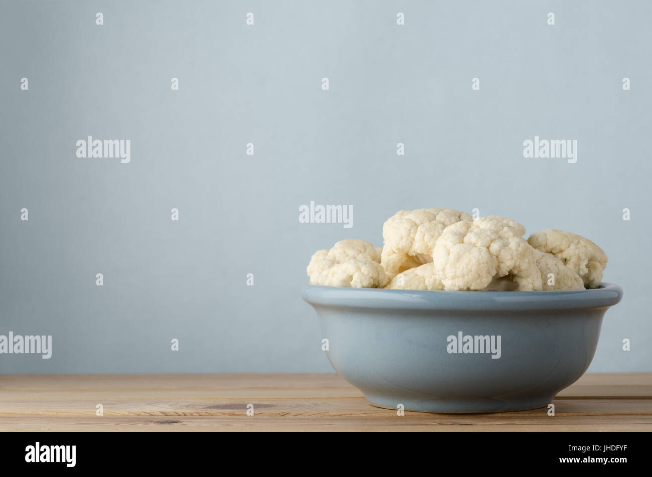 Vaso in ceramica riempito con materie prime cime di cavolfiore su luce asse di legno tabella contro il fondo azzurro con copia di spazio al di sopra e a sinistra. Foto Stock