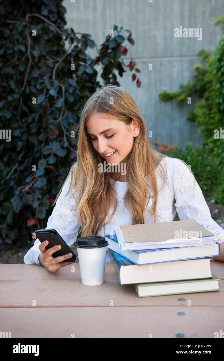 Sorridente donna giovane studente con libri e leganti distratto dal telefono mentre studiano in un area di studio di università o college campus Foto Stock