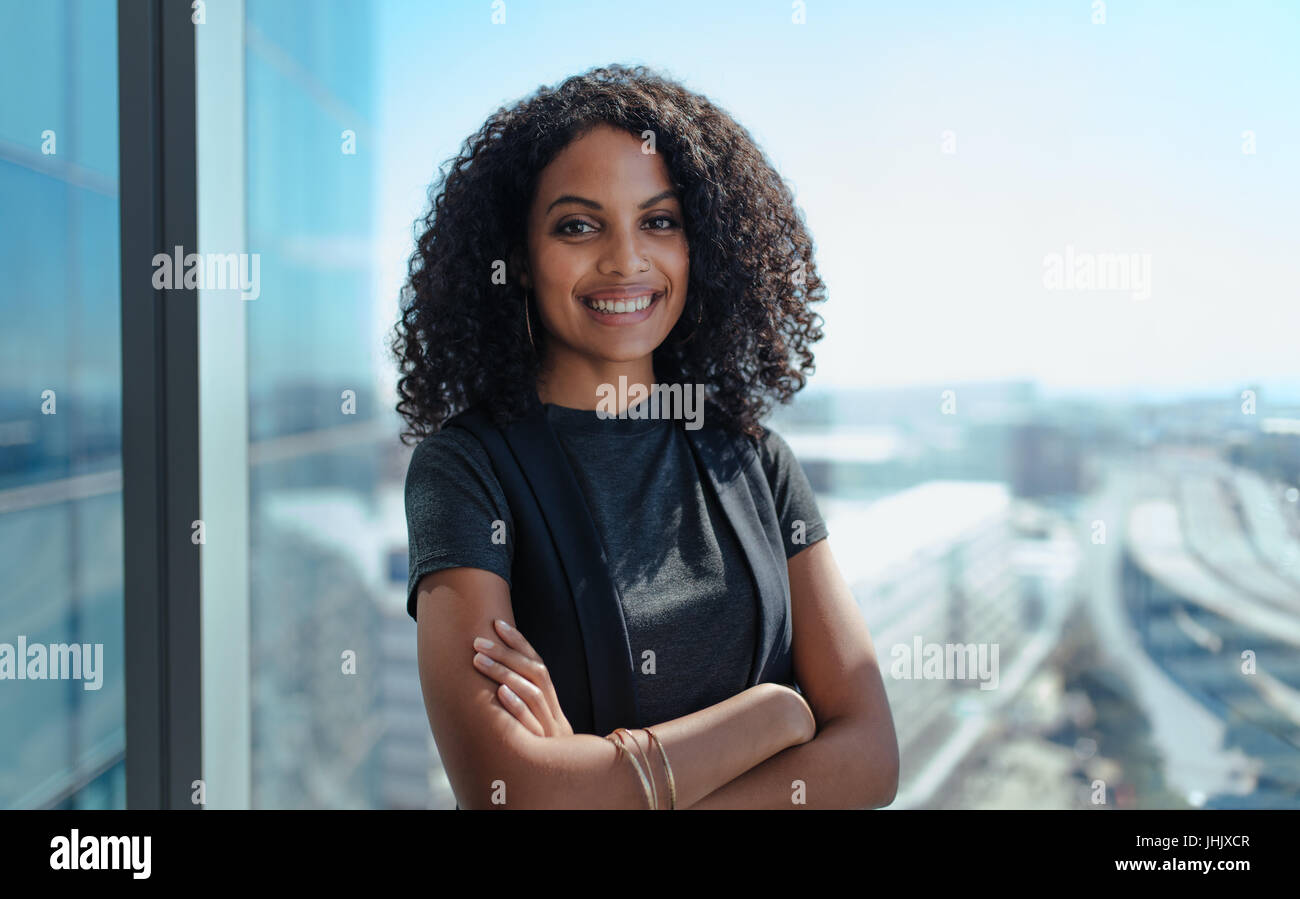 Primo piano di una imprenditrice sorridente in piedi dalla finestra del suo ufficio. Giovane donna con i capelli ricci e con le braccia incrociate in piedi in ufficio. Foto Stock