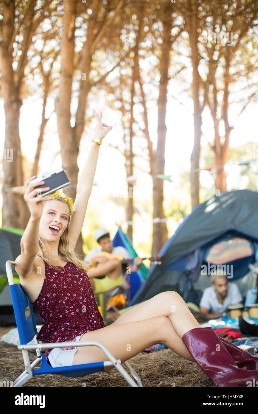 Felice giovane donna prendendo selfie mentre è seduto su una sedia al campeggio Foto Stock